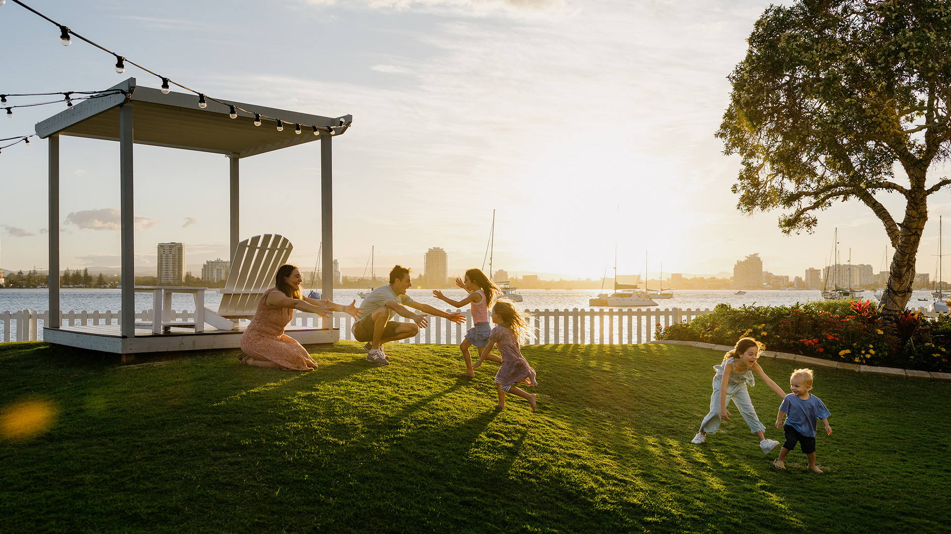 A family with young children plays and runs on a grassy lawn near a gazebo at sunset, with a waterfront city skyline and boats in the background.
