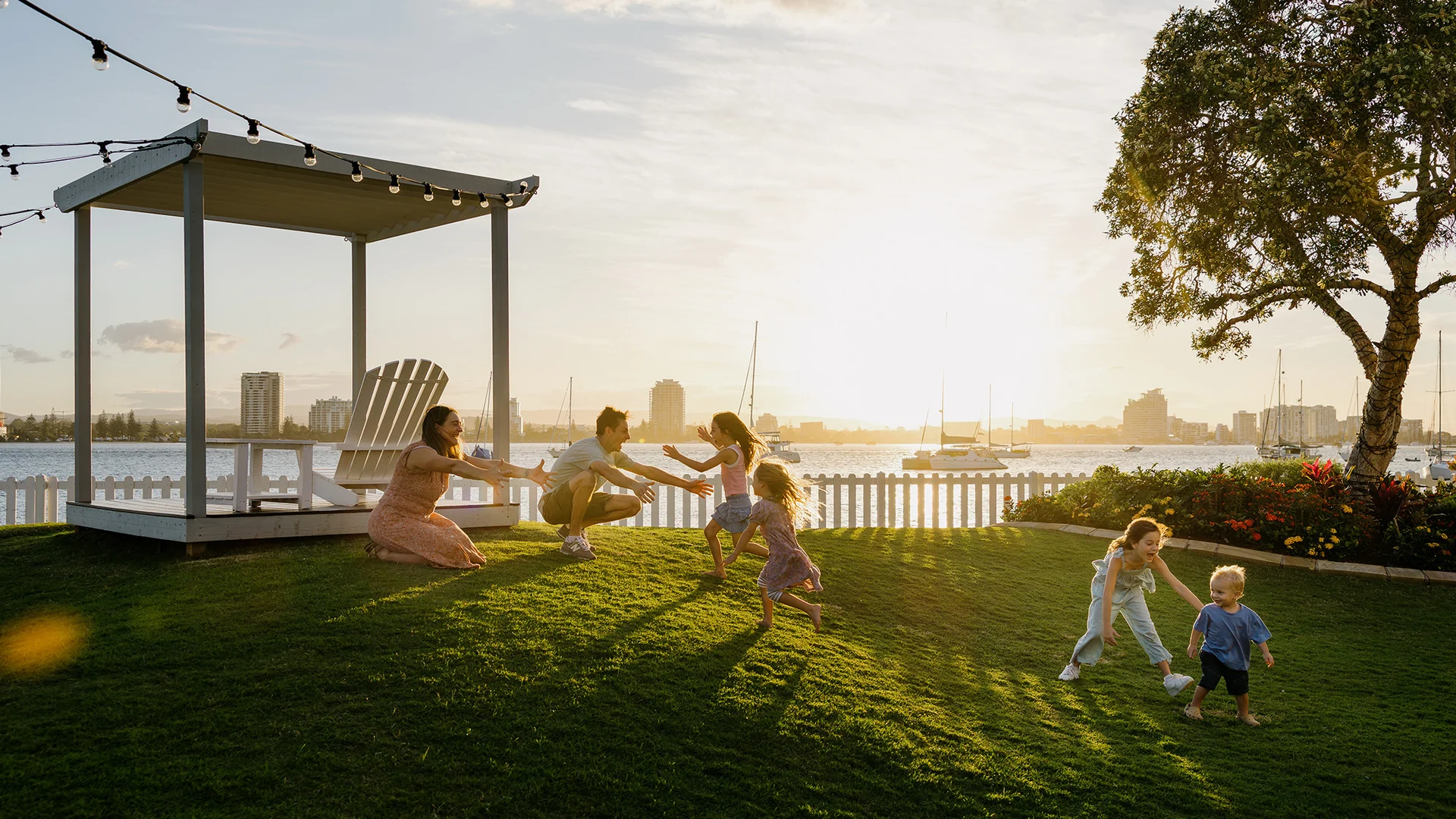 A family with young children plays and runs on a grassy lawn near a gazebo at sunset, with a waterfront city skyline and boats in the background.
