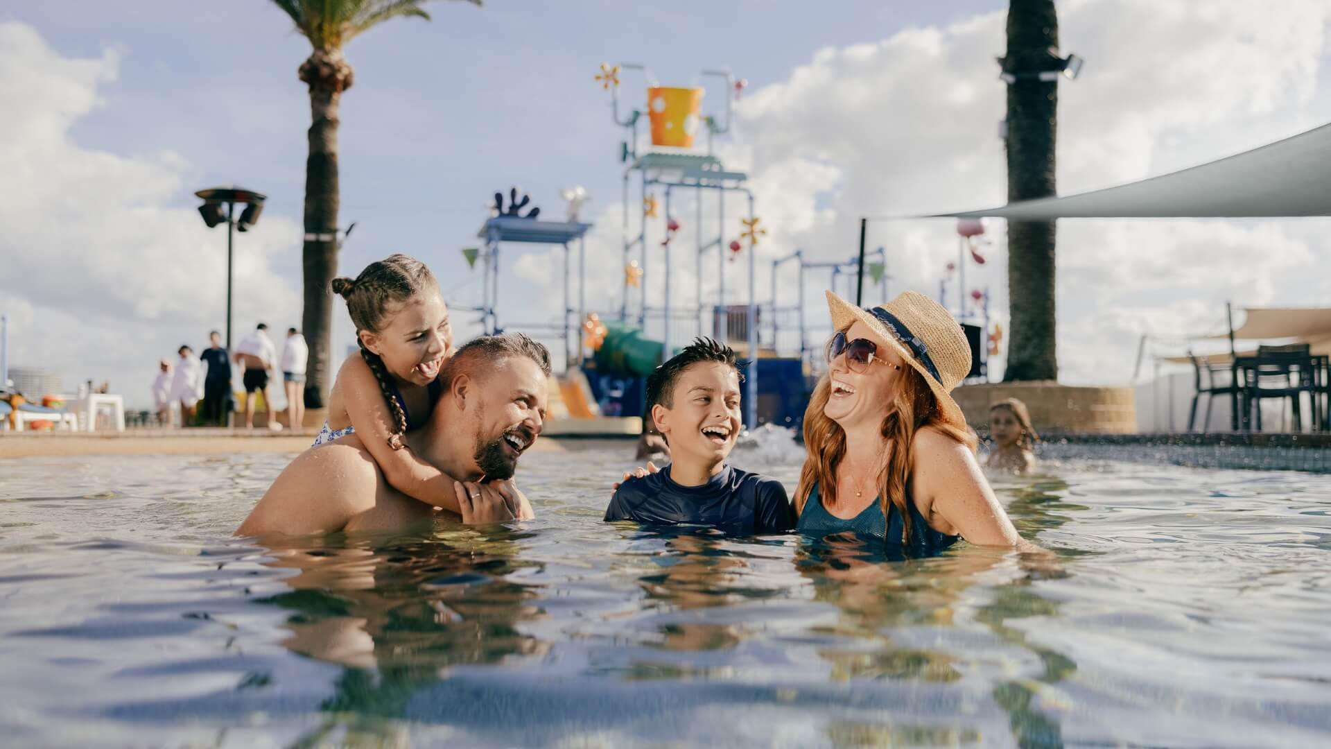 Family enjoying time in a swimming pool with a water park in the background on a sunny day.