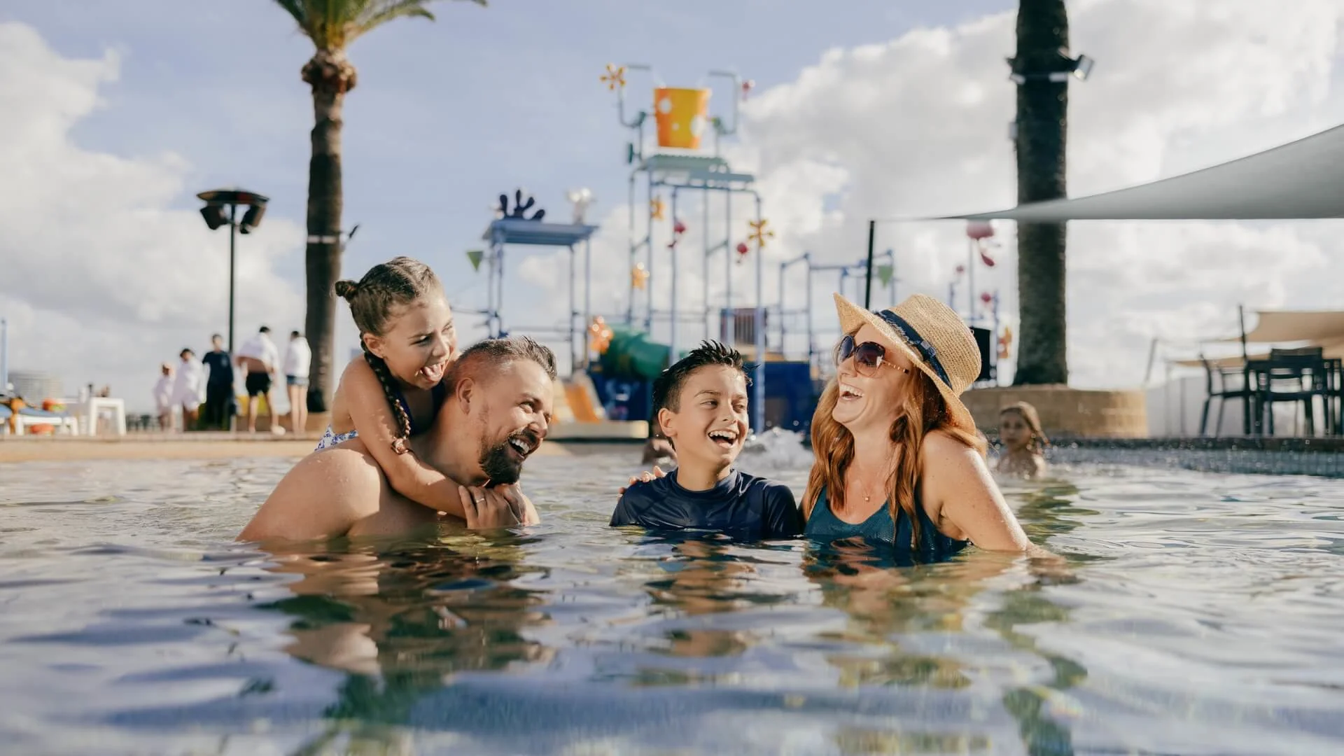 Family enjoying time in a swimming pool with a water park in the background on a sunny day.