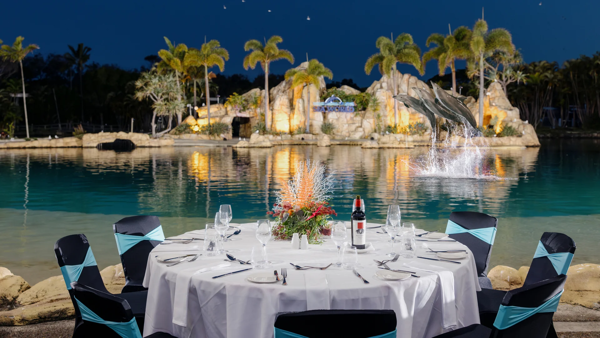 A round table set for dinner with elegant place settings and a wine bottle overlooks a lagoon with palm trees, rocks, and leaping dolphins in the background, illuminated at dusk.