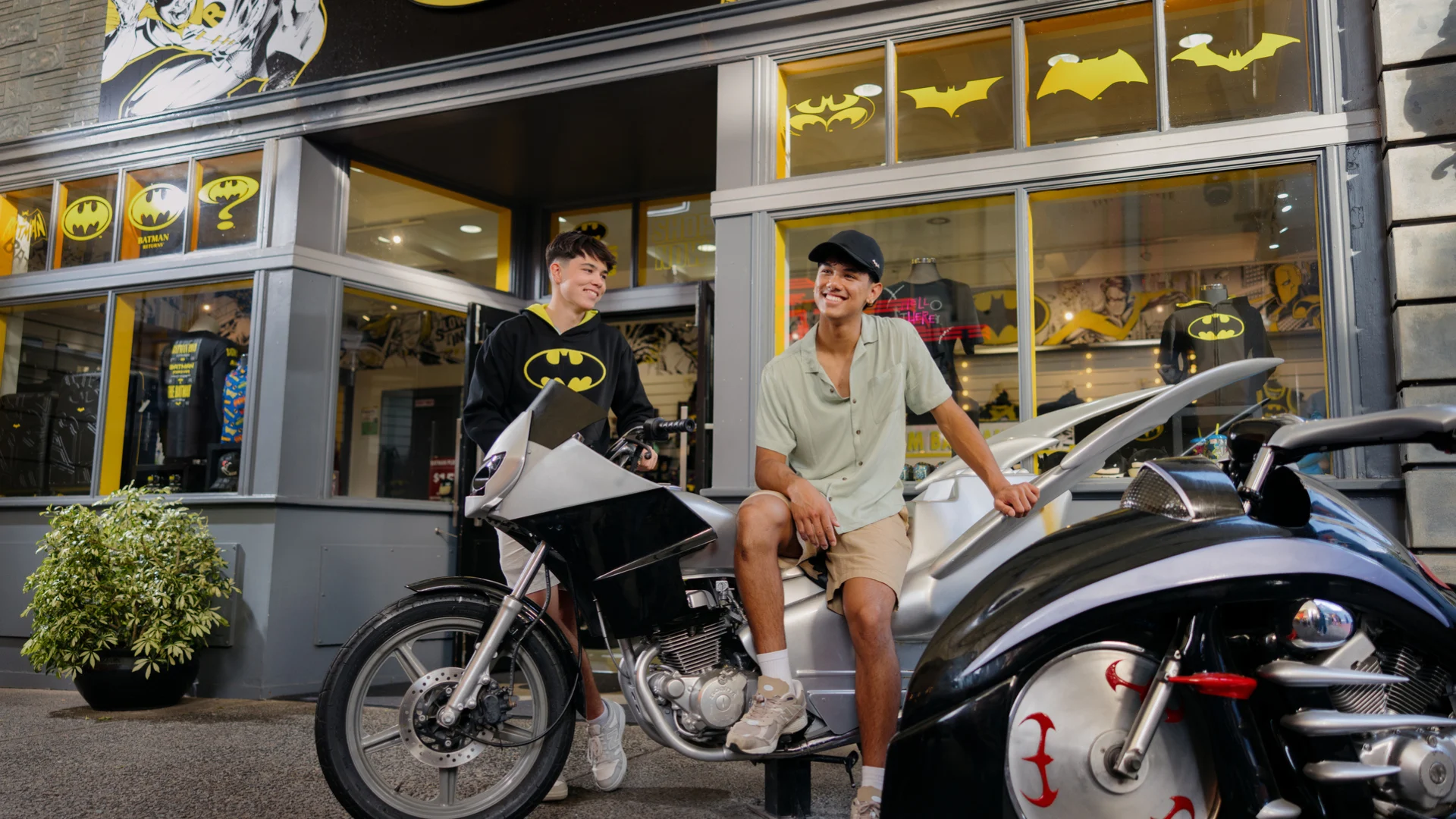 test Two people sit on a motorcycle and car in front of a Batman-themed store, smiling and conversing.