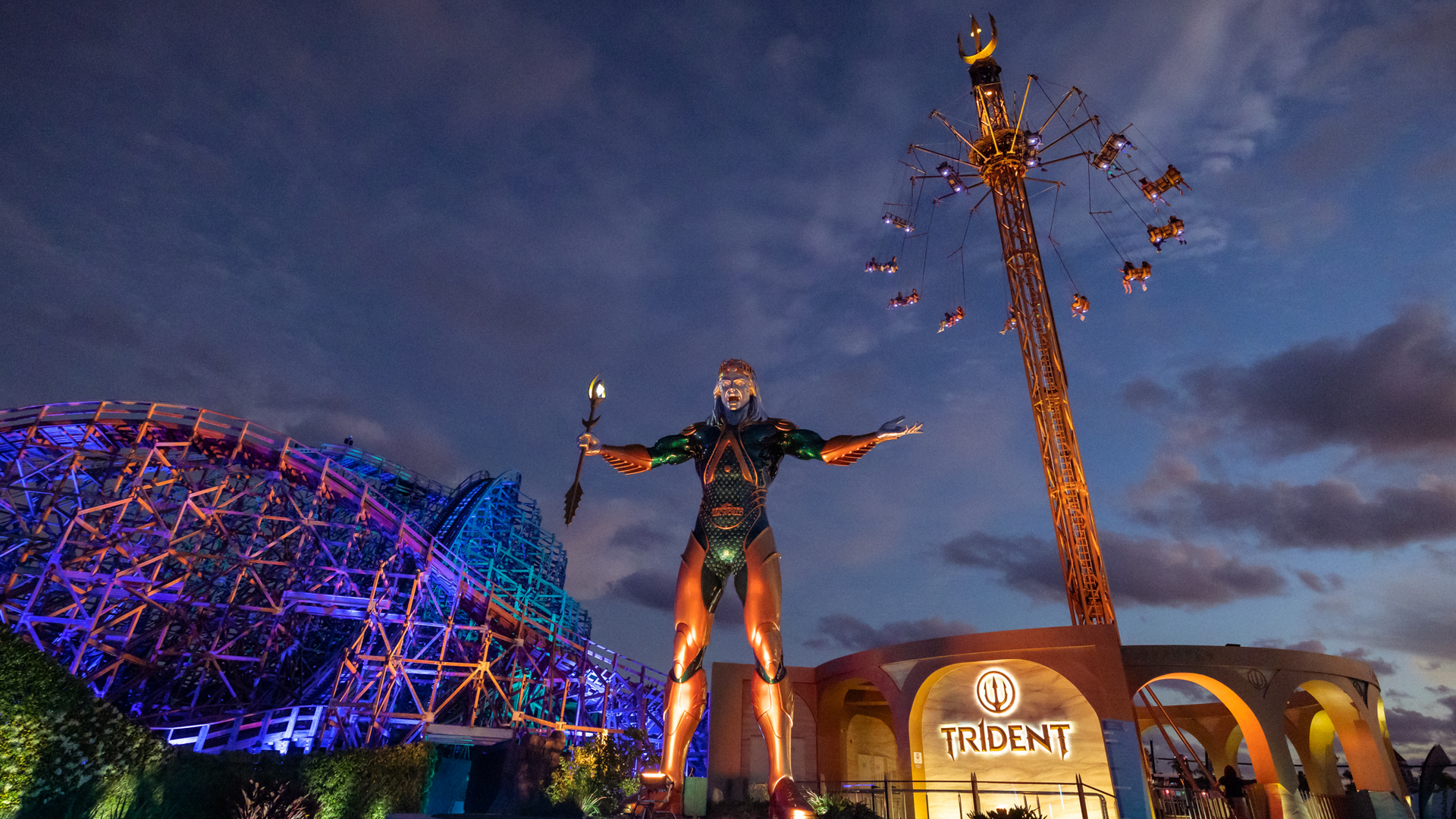A towering statue of Poseidon stands at the entrance of the Trident amusement ride, with riders spinning high in the air and a colorful roller coaster illuminated in the background at dusk.