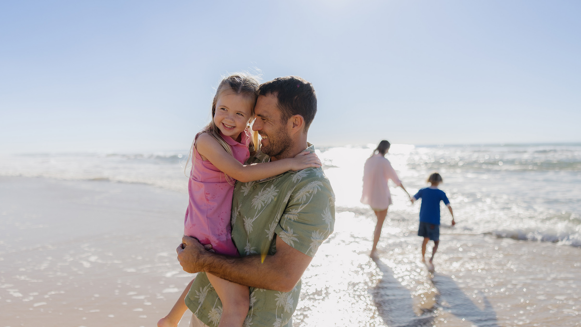 A man carries a smiling young girl on a sunny beach. In the background, a woman and a child walk along the shoreline near the water. The scene is bright and cheerful with clear blue skies.