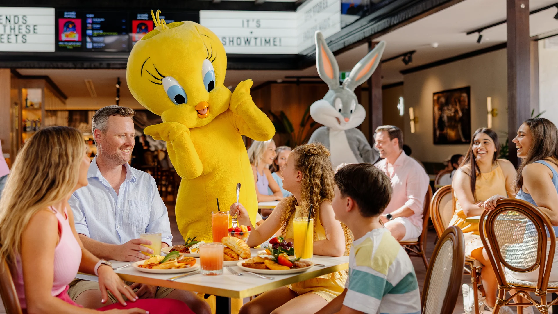 A family enjoys a meal at a restaurant while people in Tweety Bird and Bugs Bunny costumes interact with children. Everyone is smiling and the atmosphere is bright and cheerful.