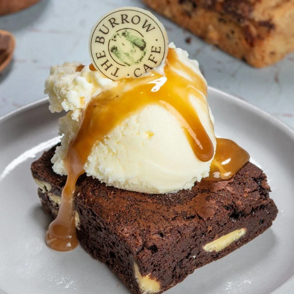 A brownie topped with a scoop of vanilla ice cream, caramel sauce, and a round "The Burrow Cafe" label on a white plate.
