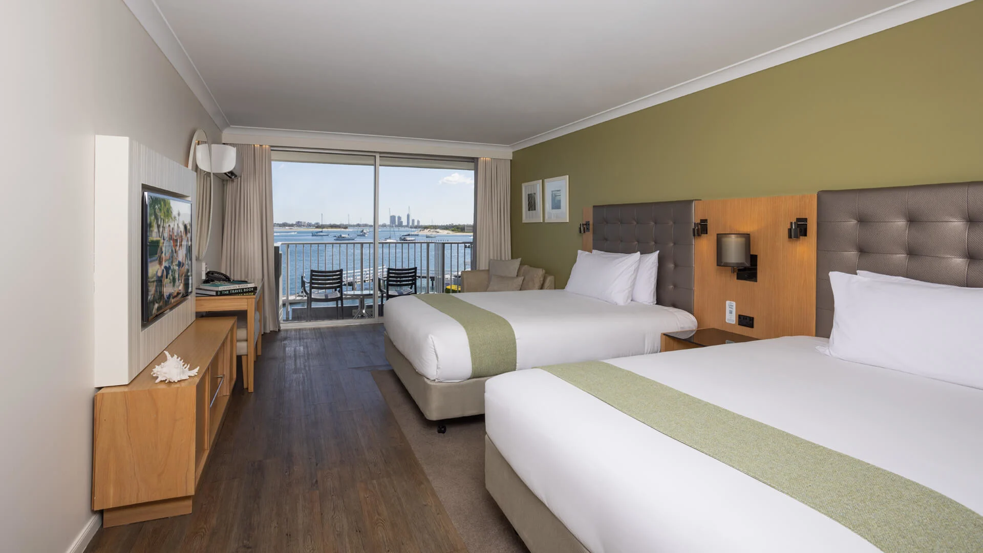 Hotel room with two double beds, green accent wall, and a view of a balcony overlooking a body of water. There's a wall-mounted TV and a small seating area.