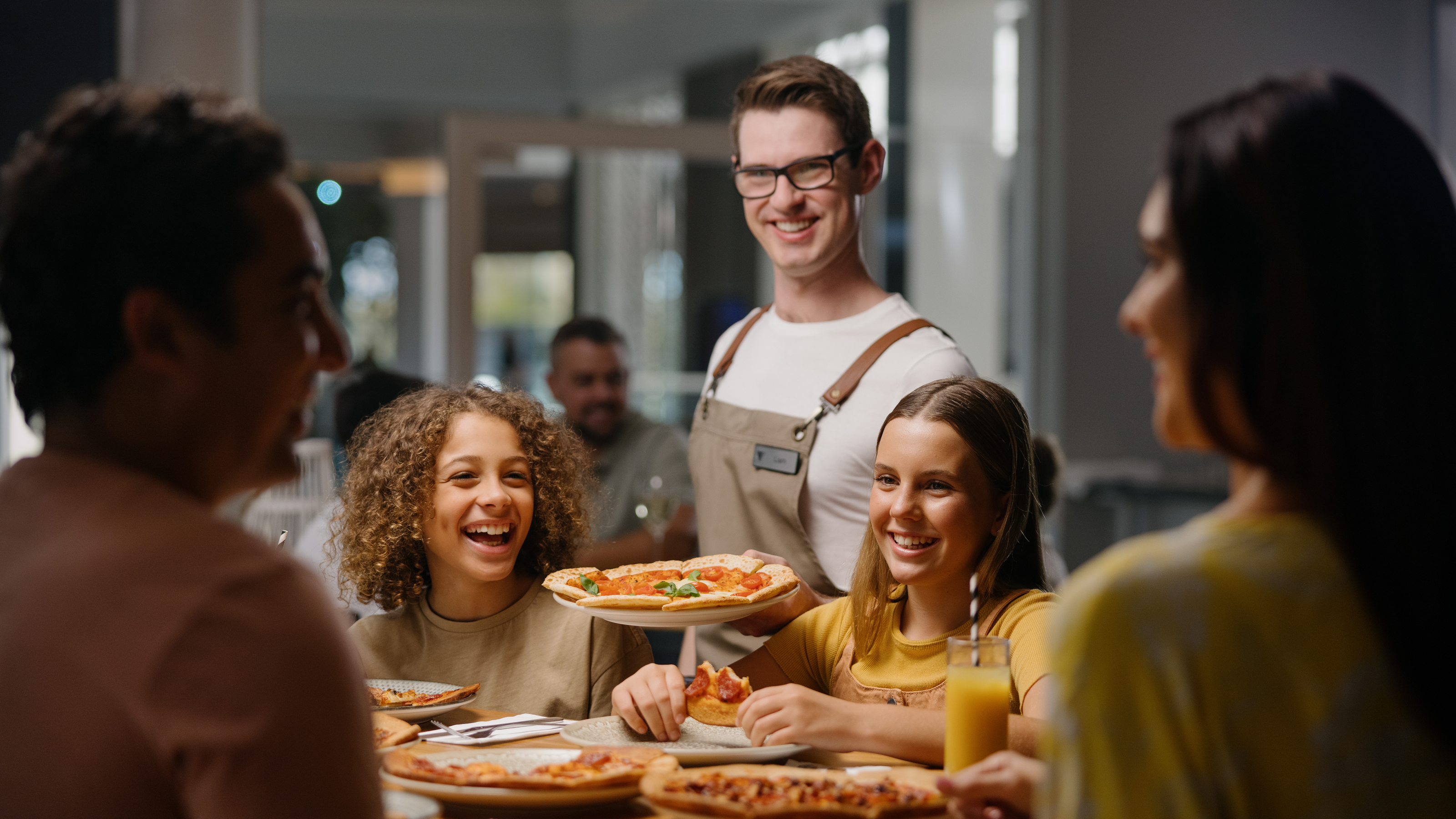 A group of people enjoying pizza at Melrose Italian Restaurant. A waiter stands nearby with a smile.
