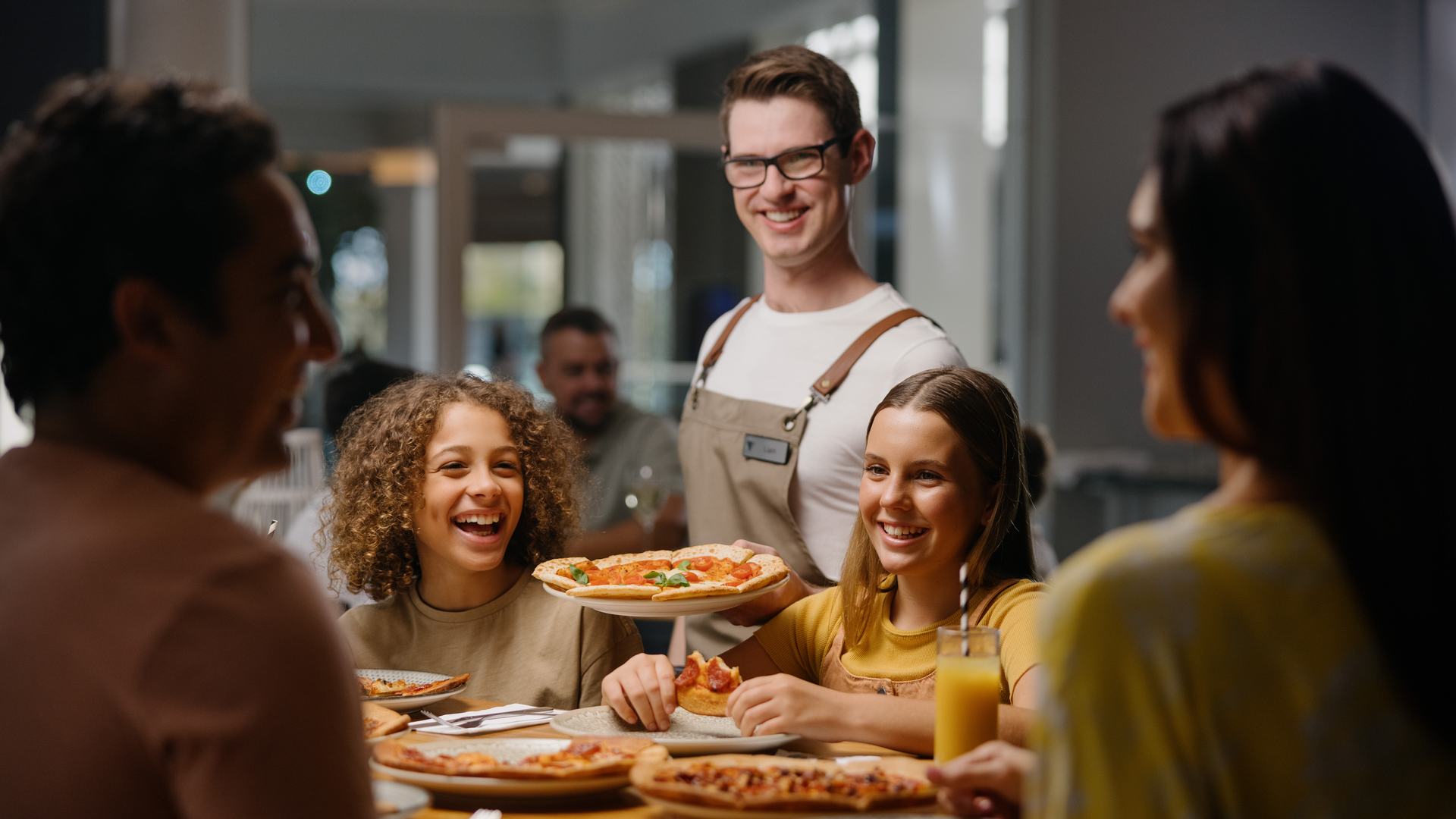 A group of people enjoying pizza at Melrose Italian Restaurant. A waiter stands nearby with a smile.