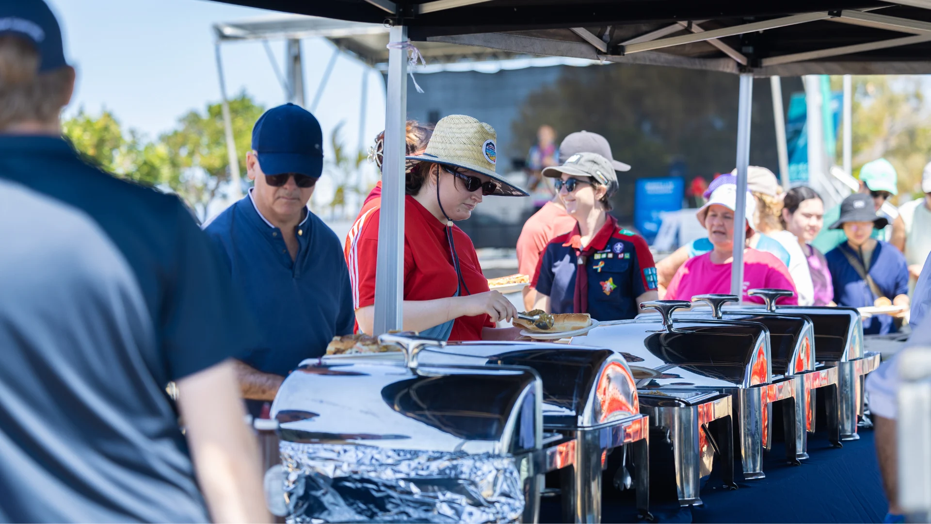 People serve themselves food from a buffet table outdoors under a canopy on a sunny day.