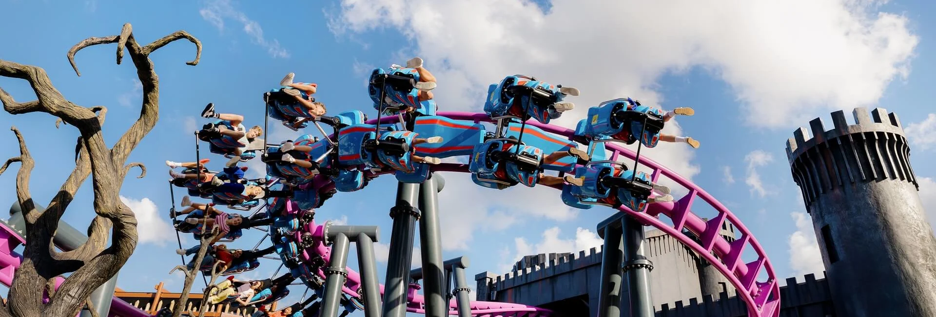 People riding a colourful looping roller coaster near a castle-themed structure under a bright blue sky.