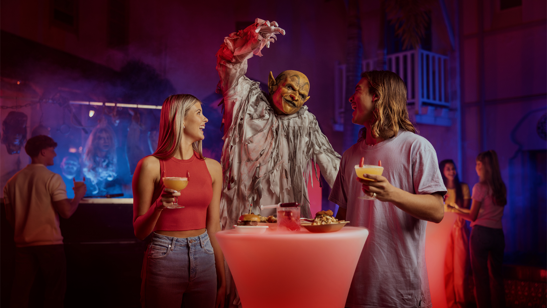 Three people at a party, two holding drinks and talking, while a person in a monster costume stands between them with raised hands. The background has colorful lighting and other guests.