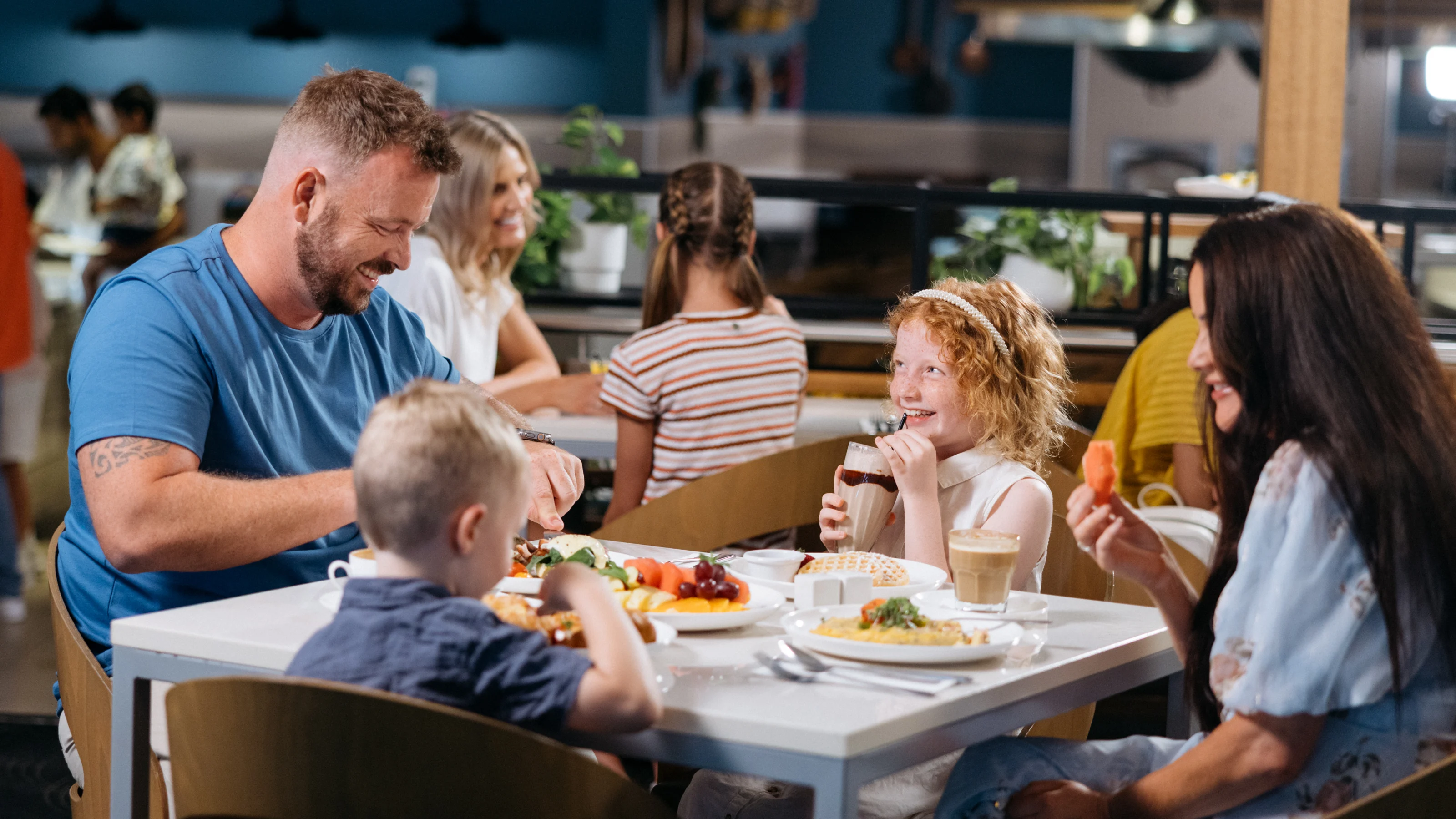 A family of four sits at a table in a restaurant, eating and smiling. The table has plates of food and drinks. Other diners are visible in the background.