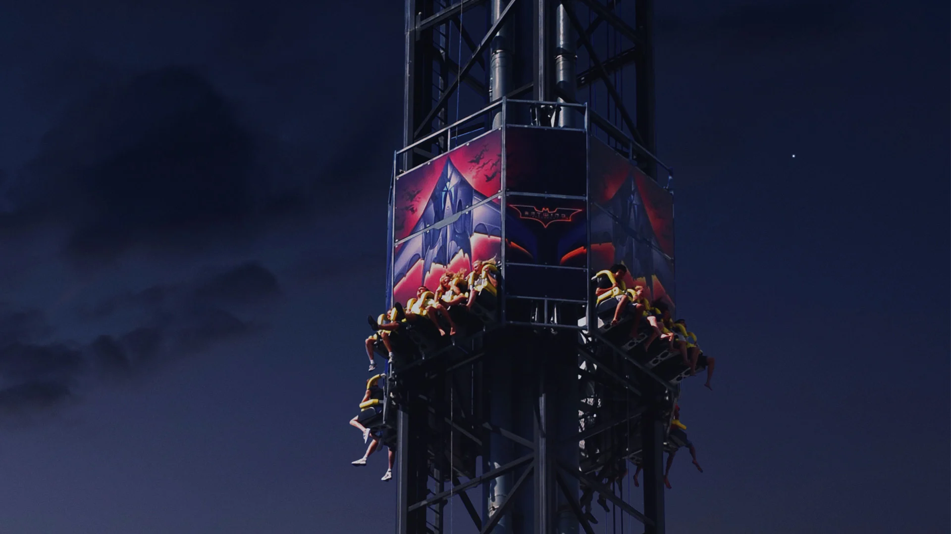 A group of people sit on a drop tower amusement park ride themed with Batman graphics, suspended high against a dark evening sky.