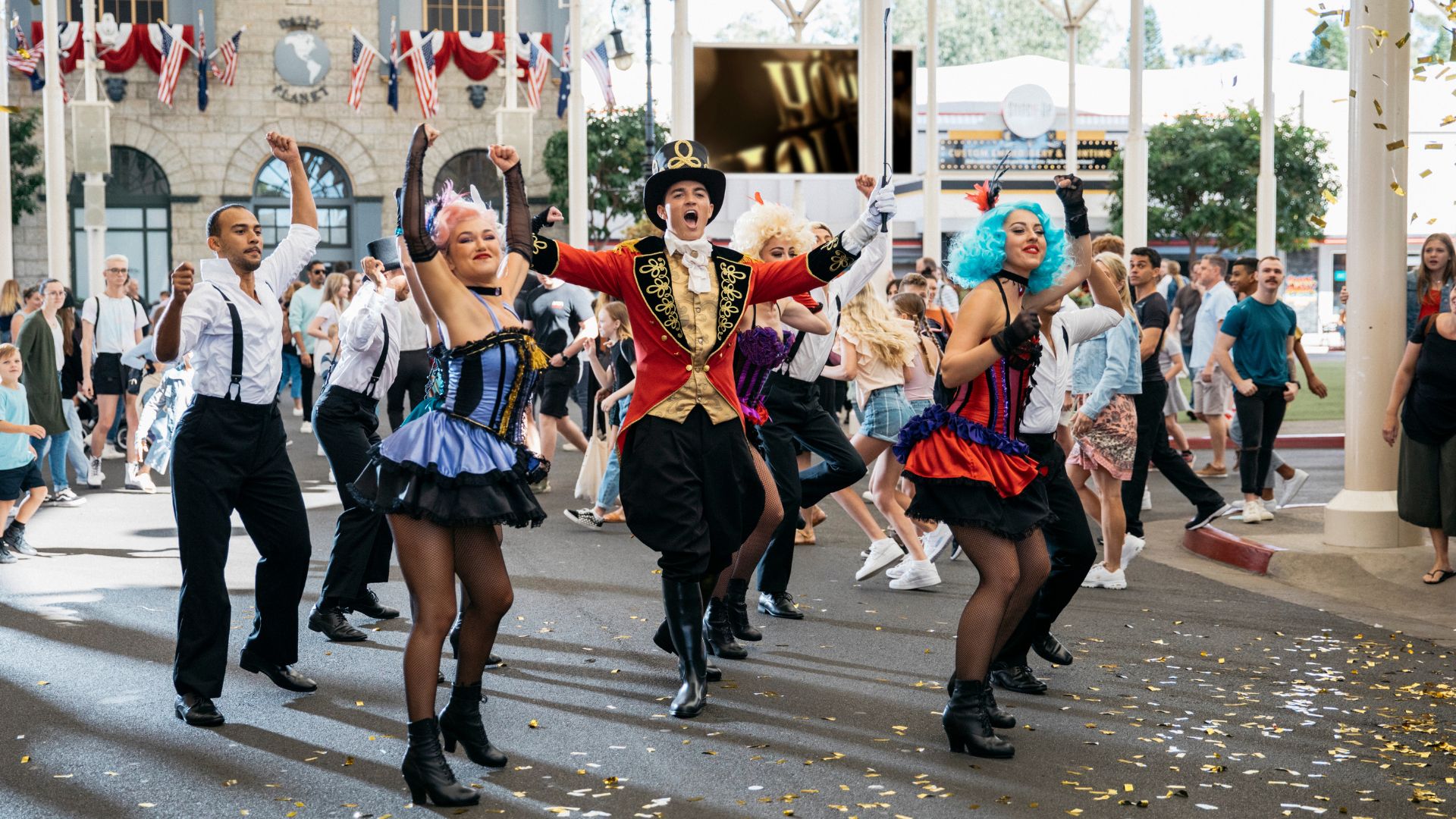A lively street performance with dancers in colorful costumes, including a person in a red ringmaster outfit and two in blue and purple dresses, entertain a crowd of people outdoors during the day.