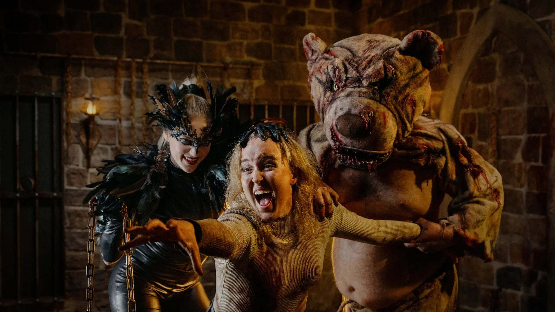 Three people in costume: one in a black feathered outfit, another in a straitjacket reaching out and screaming, and a third in a grotesque, monstrous bear suit, all in a dim, stone-walled room.