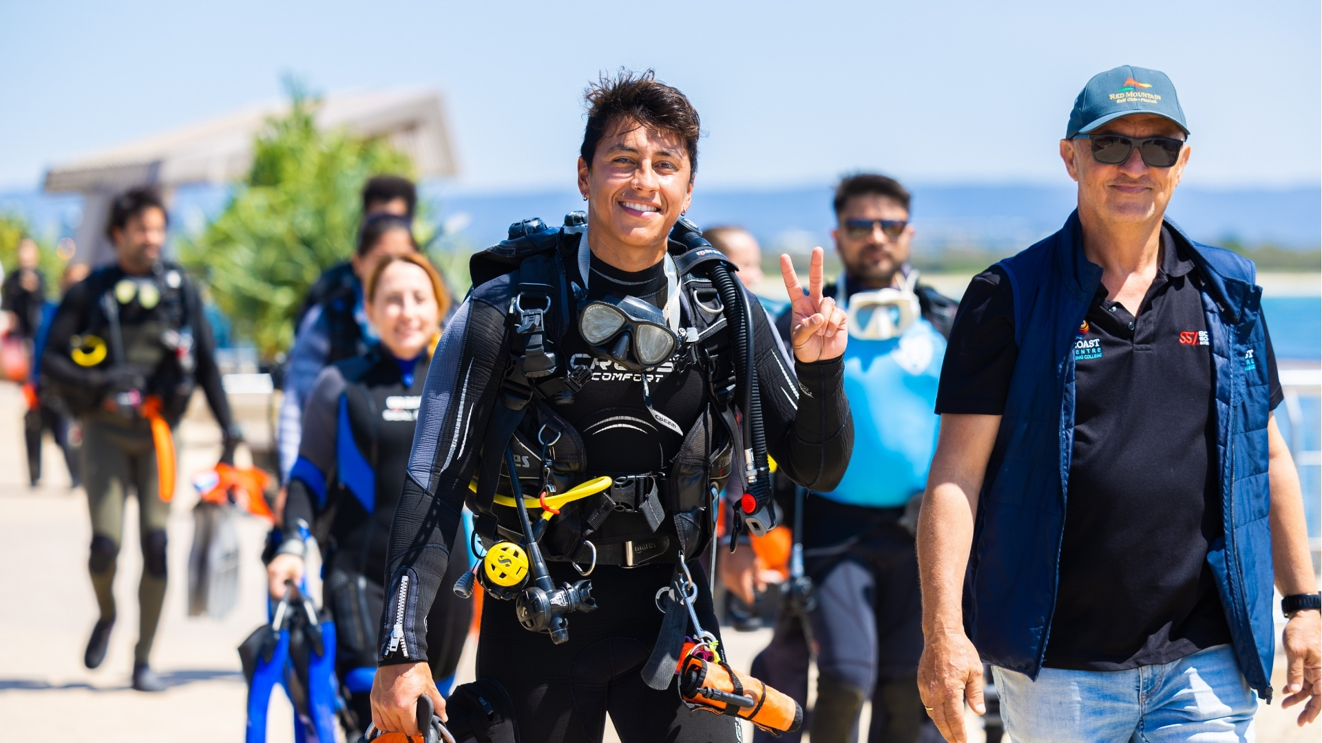 A group of people in wetsuits and scuba gear walk outdoors under a sunny sky. The person in front smiles and flashes a peace sign, while others follow behind, some carrying equipment. A man in a cap walks beside them.