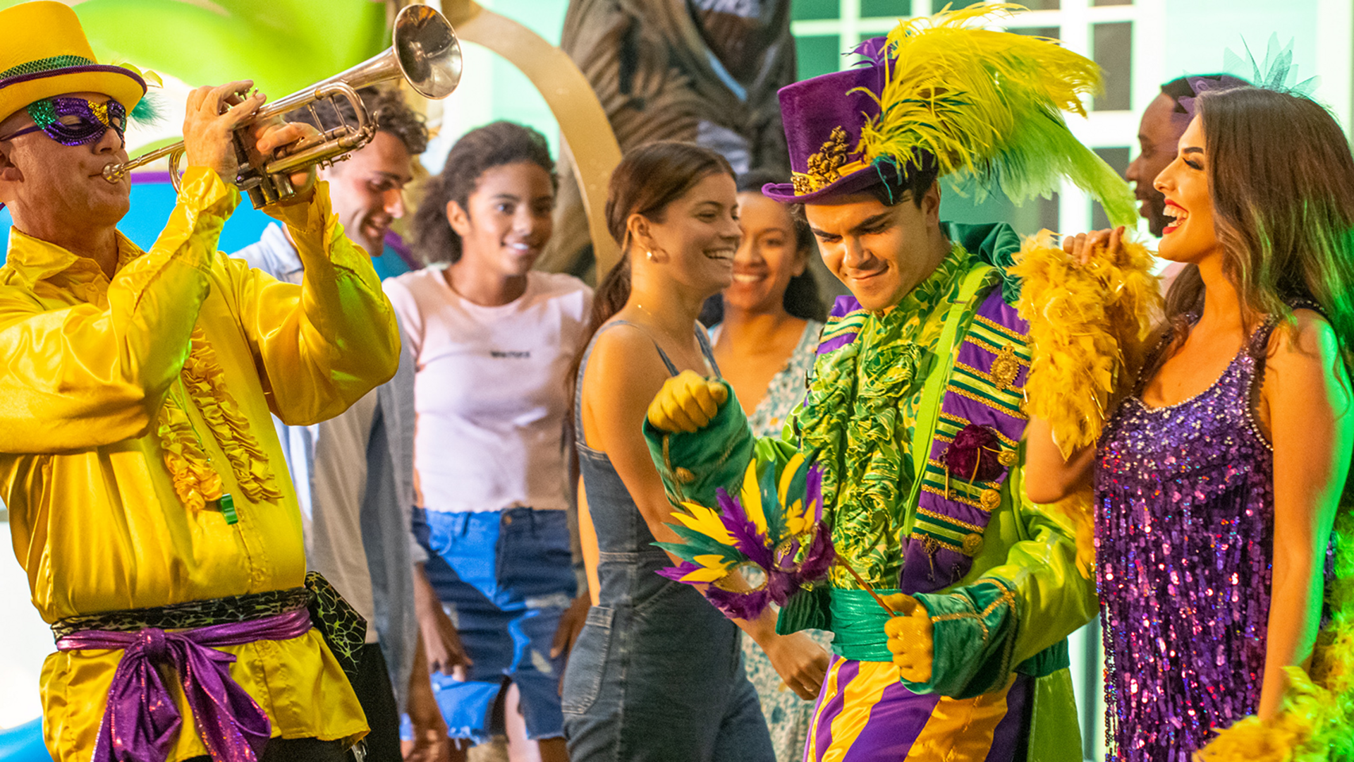 A lively group celebrates Mardi Gras; a trumpeter in bright yellow plays music as people in colorful costumes and feathered hats dance and smile, with festive decorations in the background.