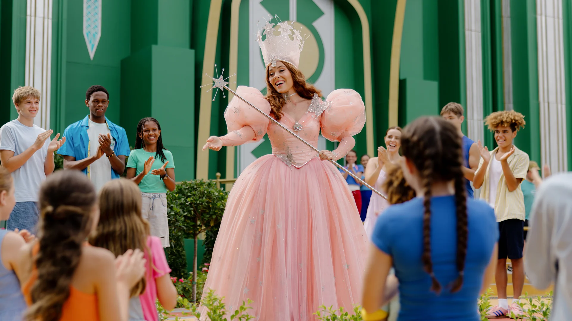 A woman dressed as a fairy tale princess in a pink gown and crown smiles and gestures with a wand while surrounded by children and teens who are watching and clapping outside a green building.