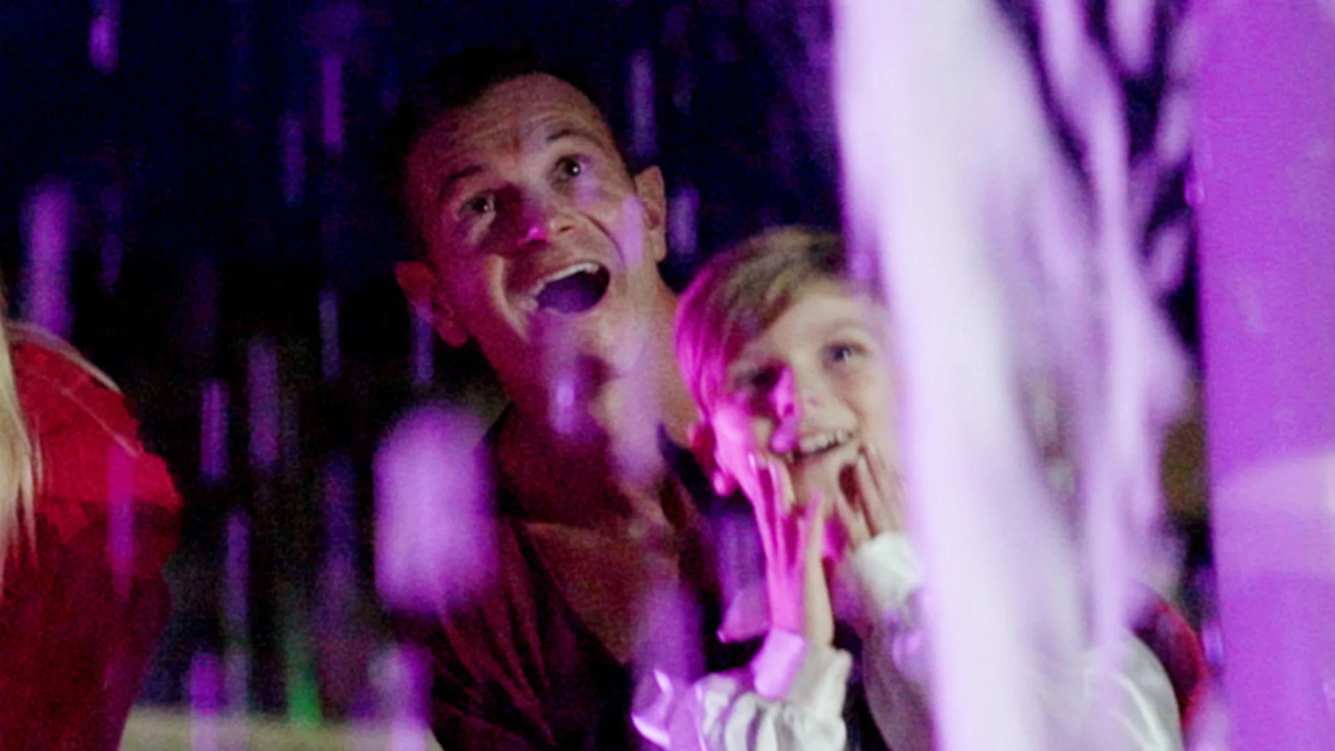 An adult and a child stand close together, gazing upward with wide smiles and expressions of awe as they are illuminated by colorful, purple-tinted lights.
