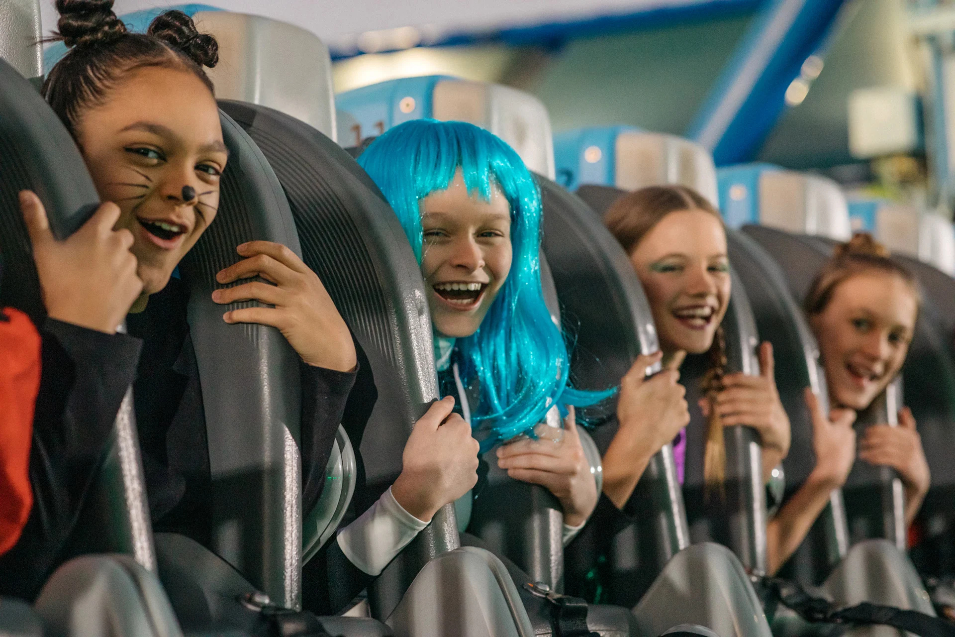 Four children in costumes sit side by side on an amusement park ride, smiling and excited. One child has a painted cat face, and another wears a bright blue wig.