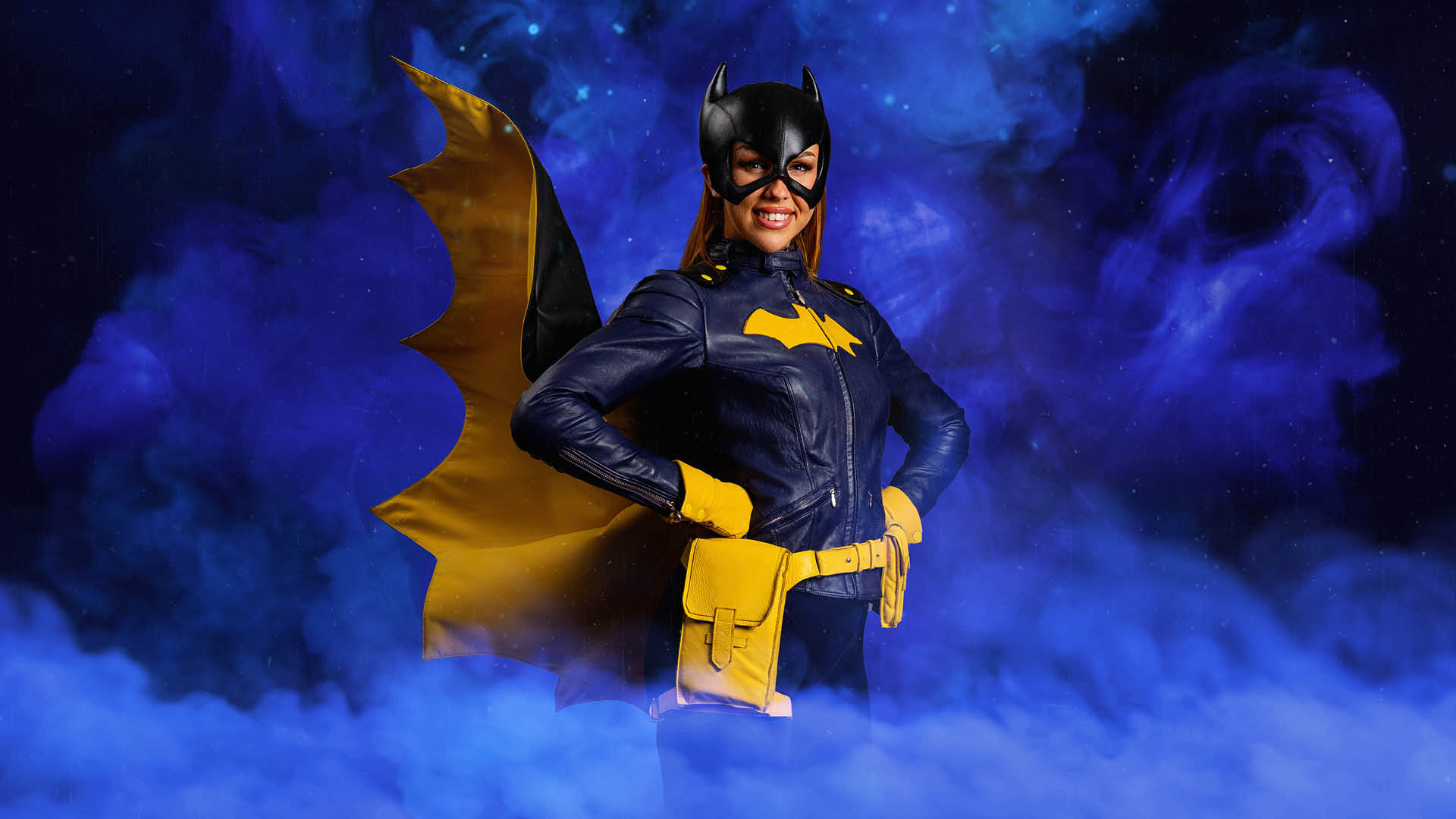 A woman dressed as Batgirl in a blue and yellow costume poses confidently with her hands on her hips against a purple smoky background.