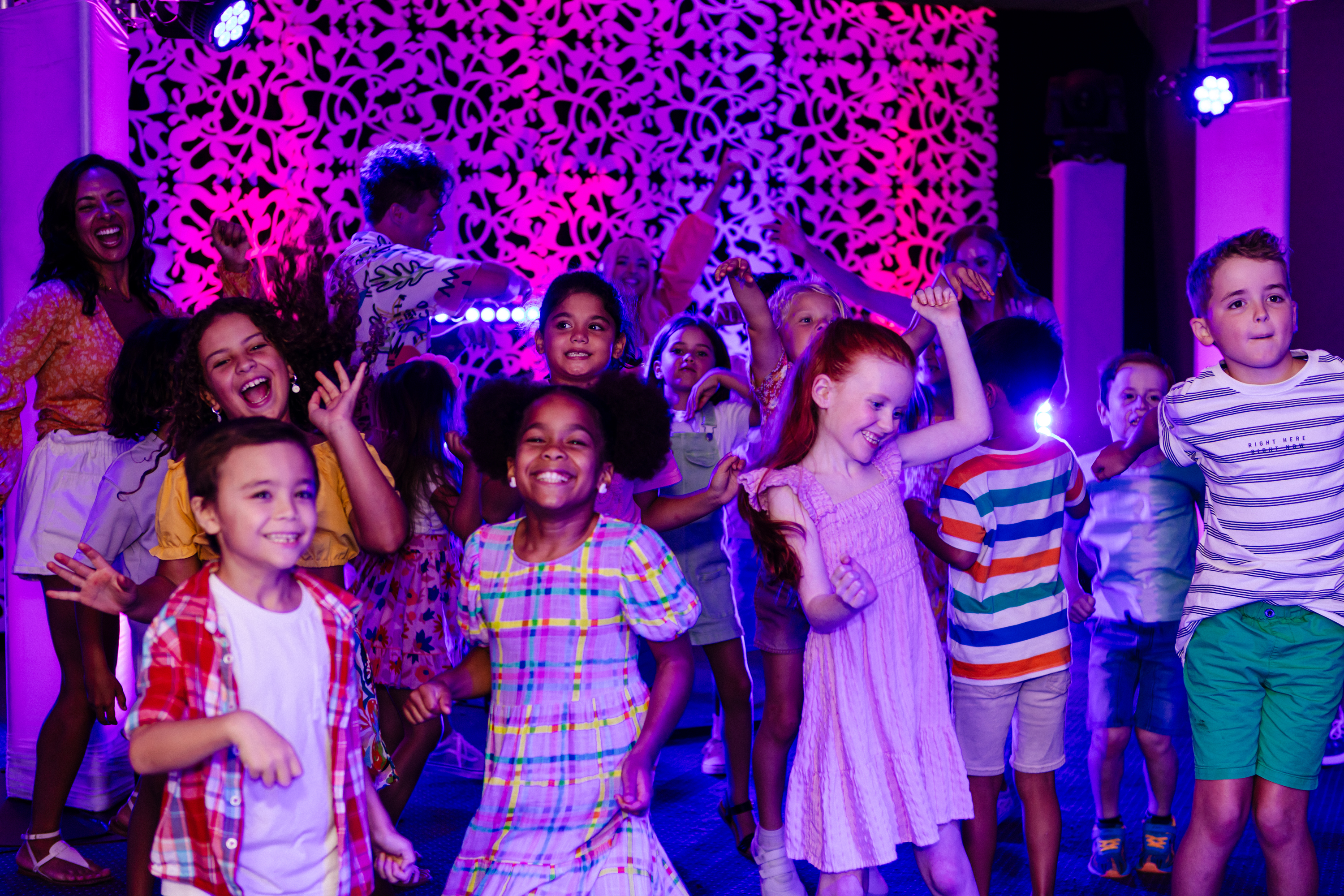 A group of children joyfully dance at a party, smiling and laughing under colorful lights, with adults in the background and a decorative pink wall behind them.