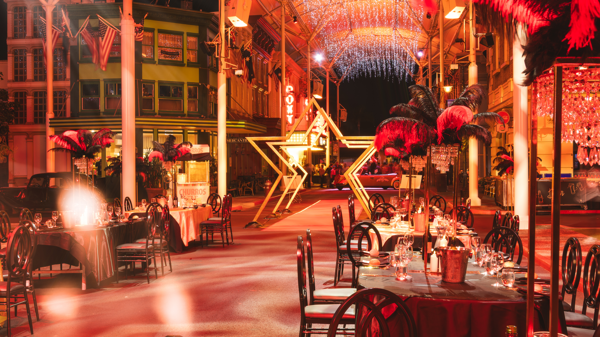 Outdoor dining area set for an event with elegant tables, black chairs, and feather centerpieces. A large illuminated star and sparkling overhead lights create a festive, glamorous atmosphere.
