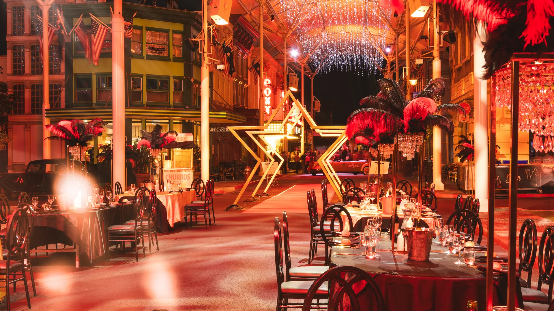 Outdoor dining area set for an event with elegant tables, black chairs, and feather centerpieces. A large illuminated star and sparkling overhead lights create a festive, glamorous atmosphere.