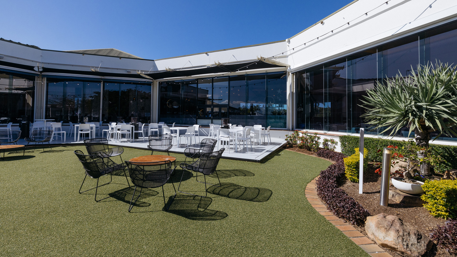 Outdoor seating area with black and white chairs on a grassy patio, adjacent to a modern building with large windows and surrounded by landscaping.