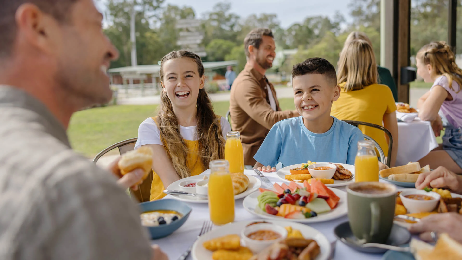 A family, including children, sit at an outdoor table enjoying breakfast foods and orange juice, with greenery in the background.