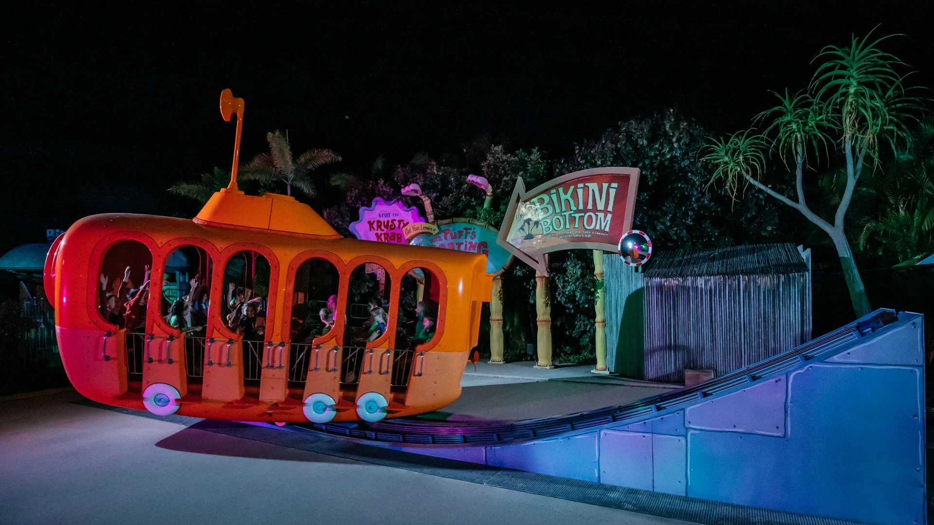 A bright orange amusement park ride shaped like a submarine is filled with people. The ride is near a sign that reads "Bikini Bottom," surrounded by tropical plants and illuminated at night.