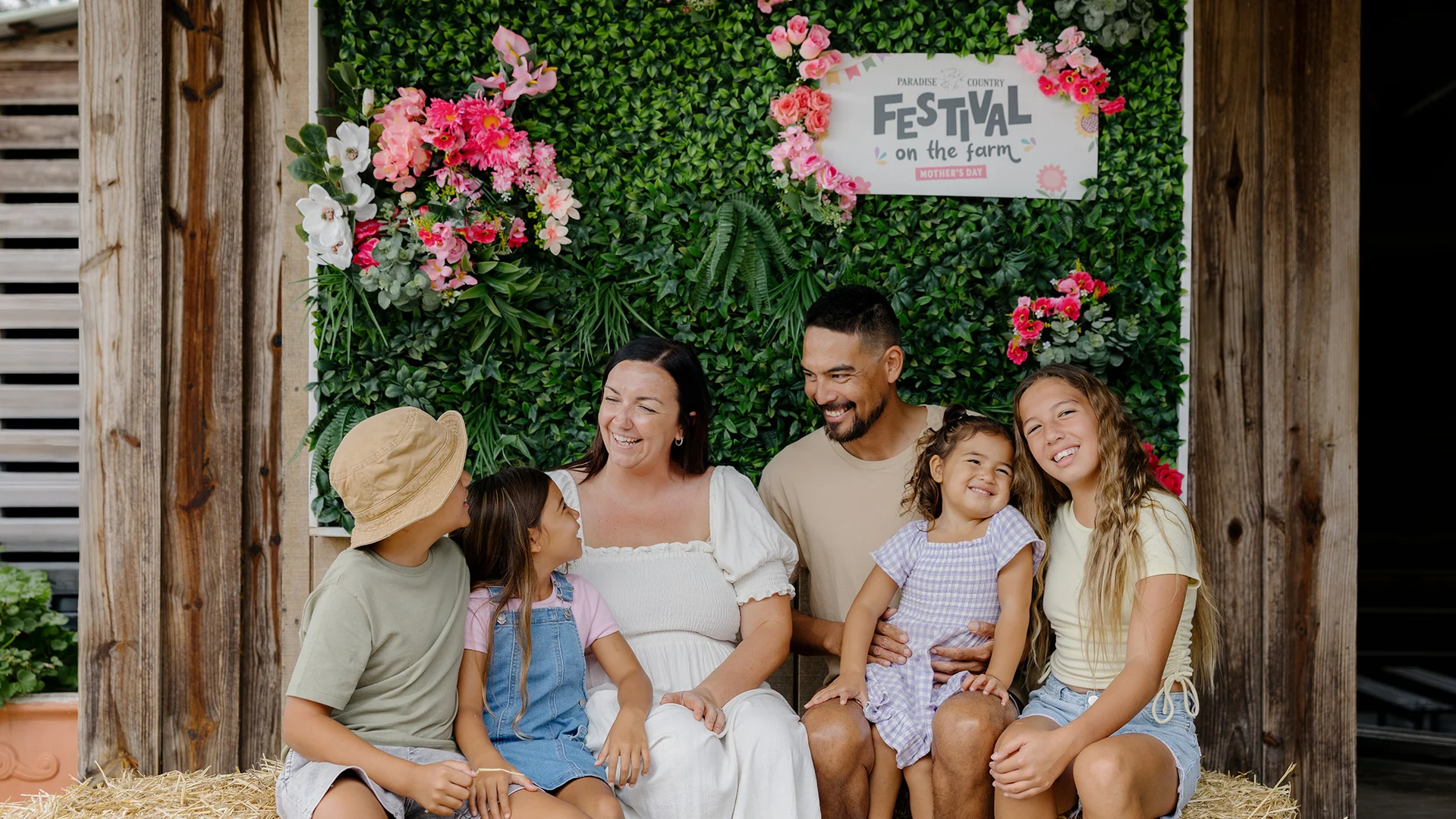 A smiling family of five sits on hay in front of a green floral wall with a “Festival on the Farm” sign. The parents and three children enjoy the festive, rustic setting together.