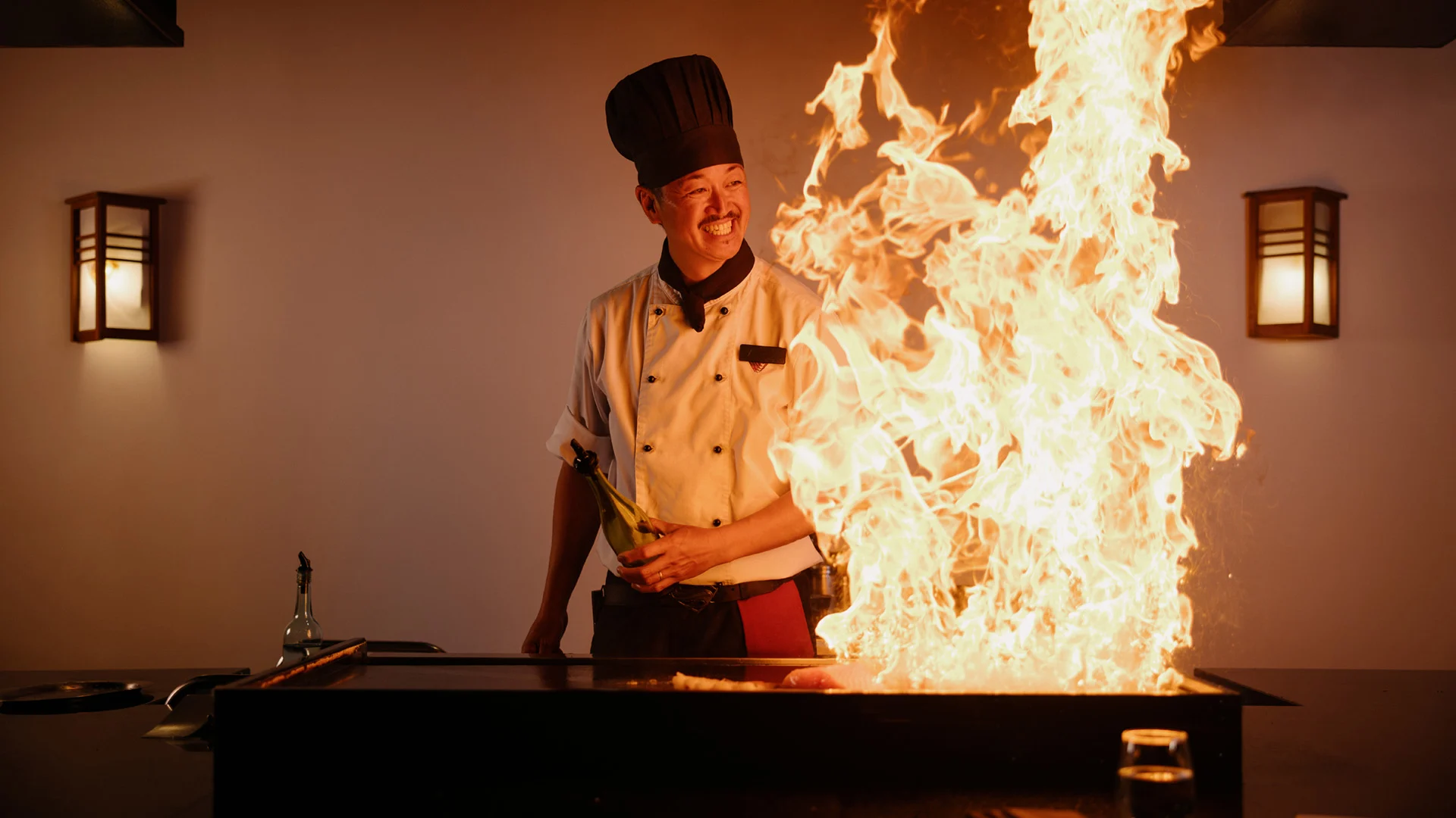 Chef in a uniform and hat smiles while cooking on a large teppanyaki grill with tall flames at Hatsuhana Japanese Restaurant, flanked by wall-mounted lights.
