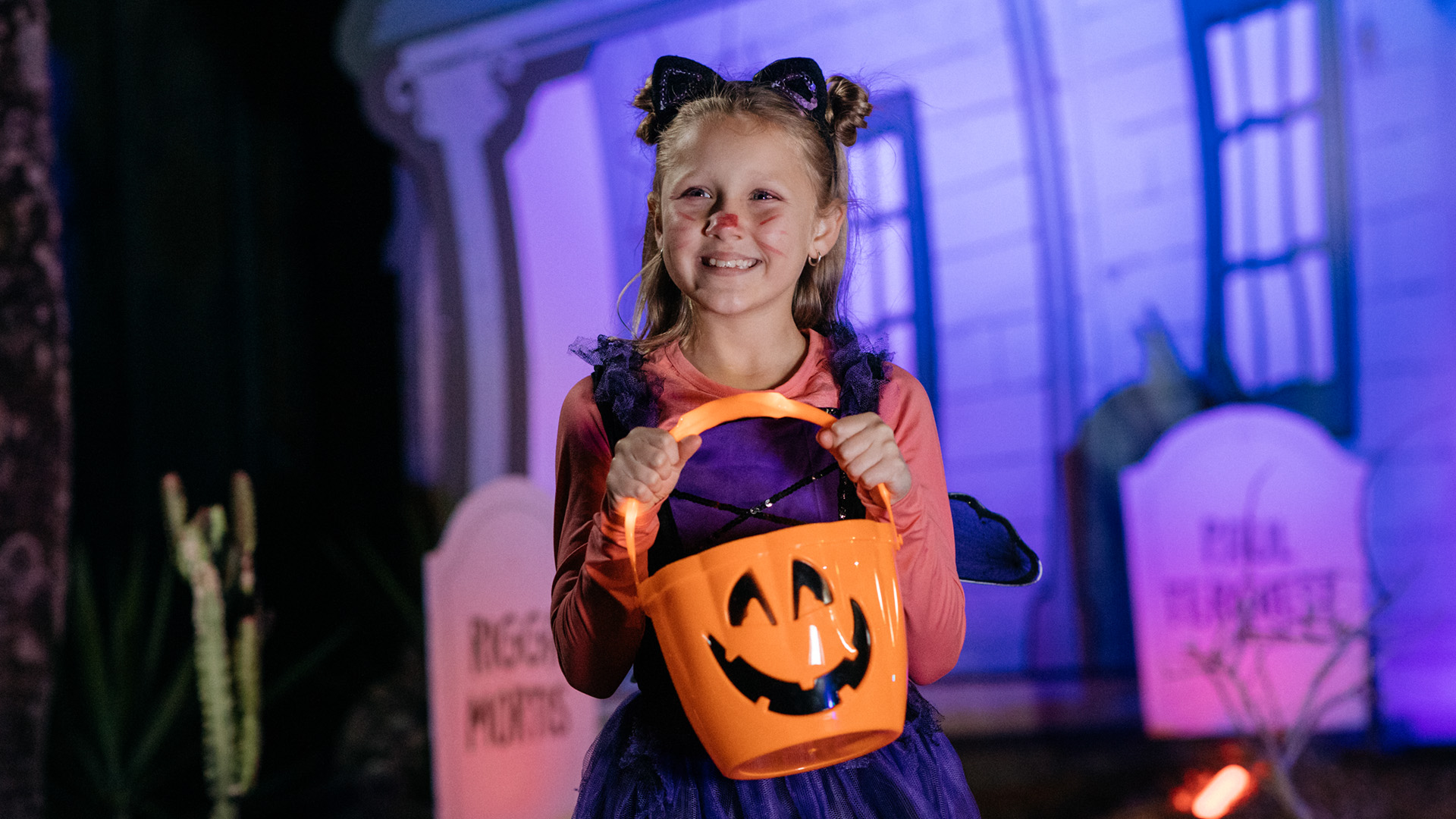 A young girl in a cat costume smiles while holding an orange pumpkin-shaped bucket. She stands in front of Halloween decorations, including tombstones and a skeleton arm, with purple and blue lighting in the background.