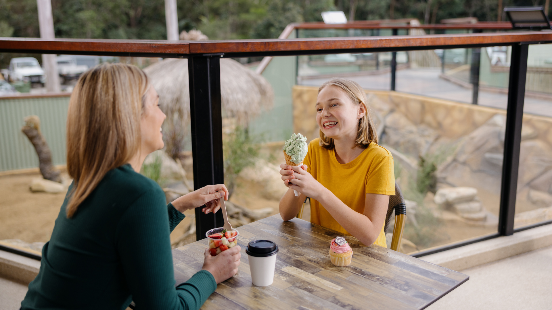 Two people sit at a table by a window, one holding an ice cream cone and smiling, the other with a coffee and a parfait, with desserts on the table.