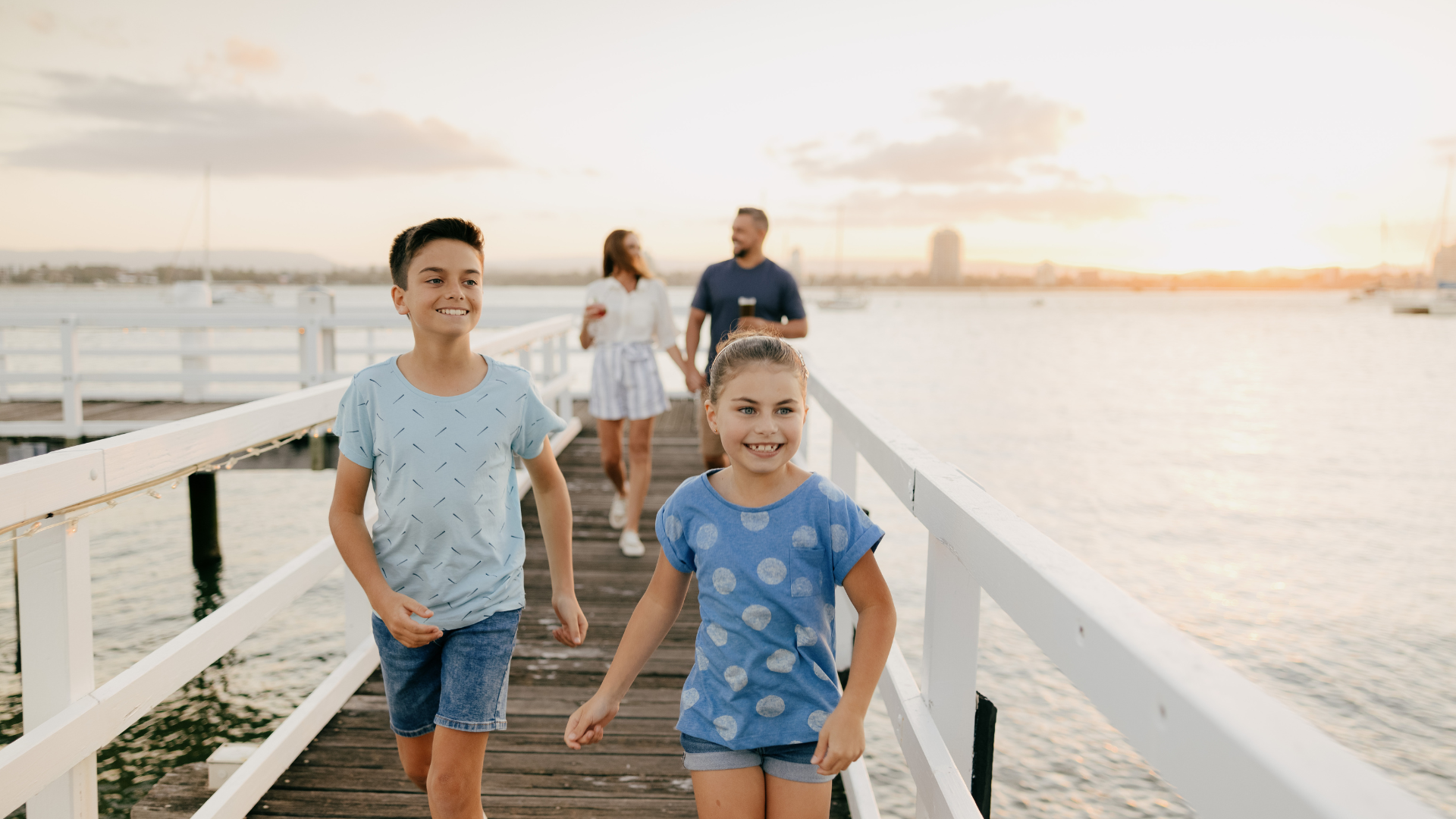 Two children in blue shirts stroll down the jetty at the Boat Shed, Sea World Resort, their laughter blending with the whispers of a sunset sky. Behind them, two adults follow, enjoying the serene view.