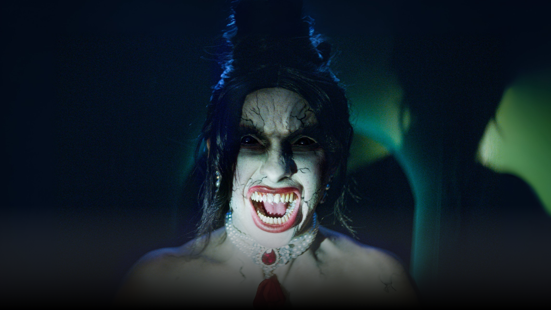 A person with a pale, ghostly face, exaggerated dark makeup, sharp fangs, and a menacing expression screams. They wear a jeweled choker necklace, with shadows and dim light in the eerie background.