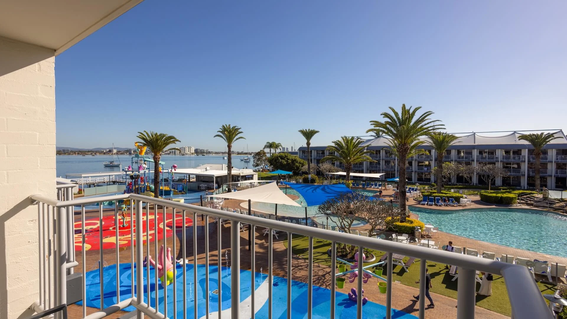 Balcony view of a resort with a large pool, palm trees, and lounge areas. A body of water and distant buildings are visible in the background under a clear sky.