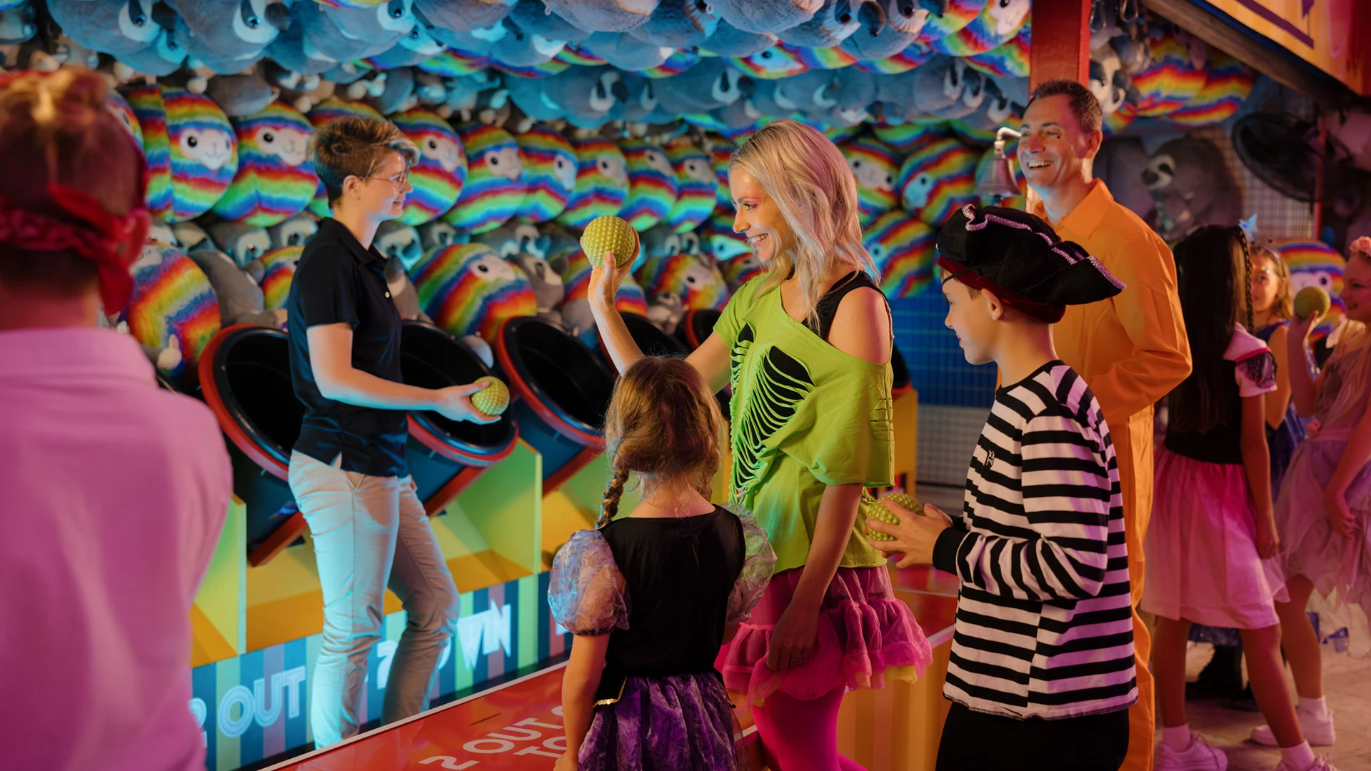 People in colorful costumes play a ball toss carnival game surrounded by plush prizes shaped like rainbow fish. An attendant watches as players throw balls, and others wait their turn, smiling and enjoying the activity.