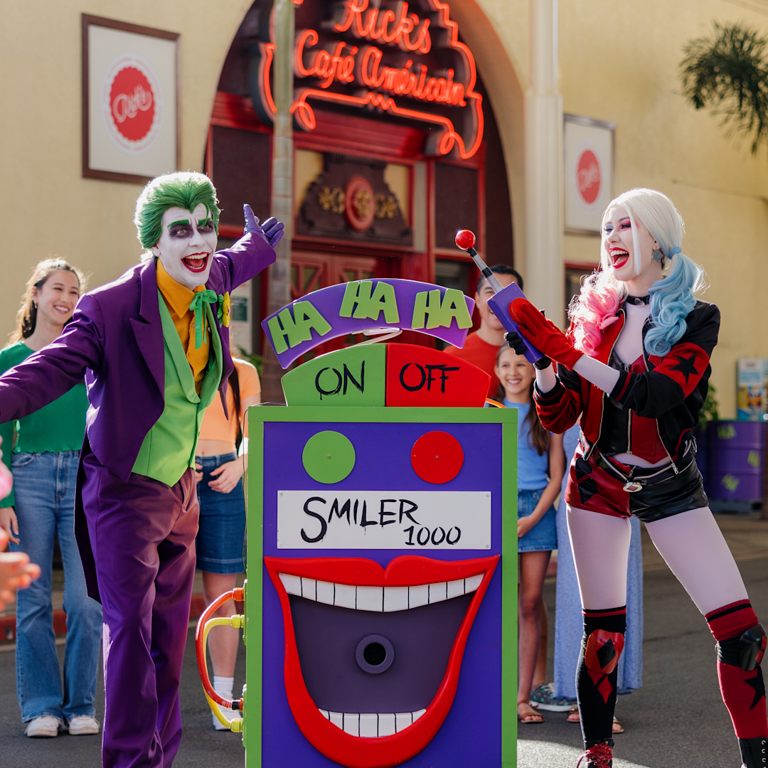 Two people dressed as the Joker and Harley Quinn stand by a large, colorful "Smiler 1000" machine, entertaining a group of onlookers outside a café.