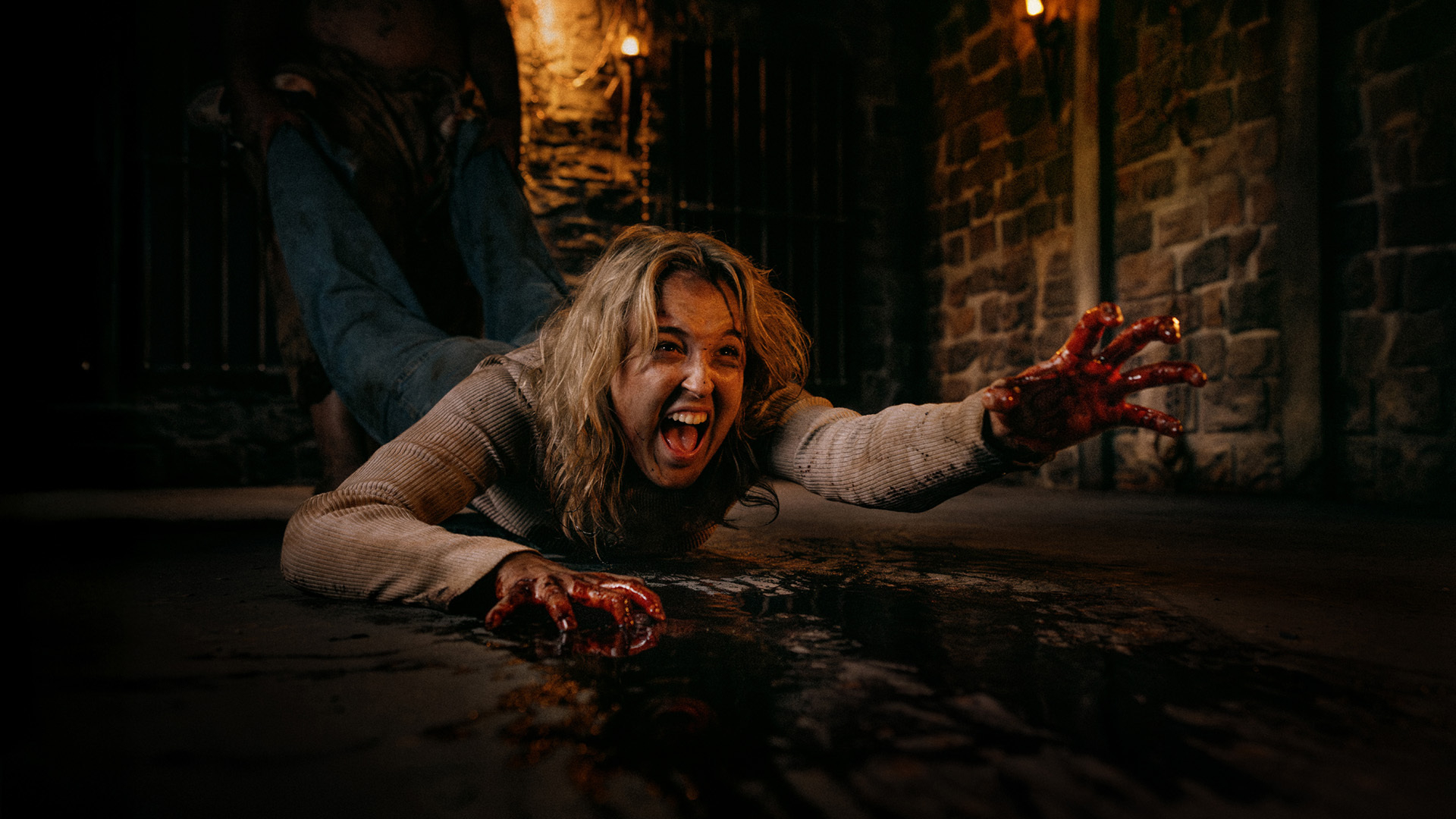 A terrified woman with bloodied hands screams and reaches forward while being dragged across a dark, stone-floored room by someone behind her. The scene is tense and dramatic with dim, eerie lighting.