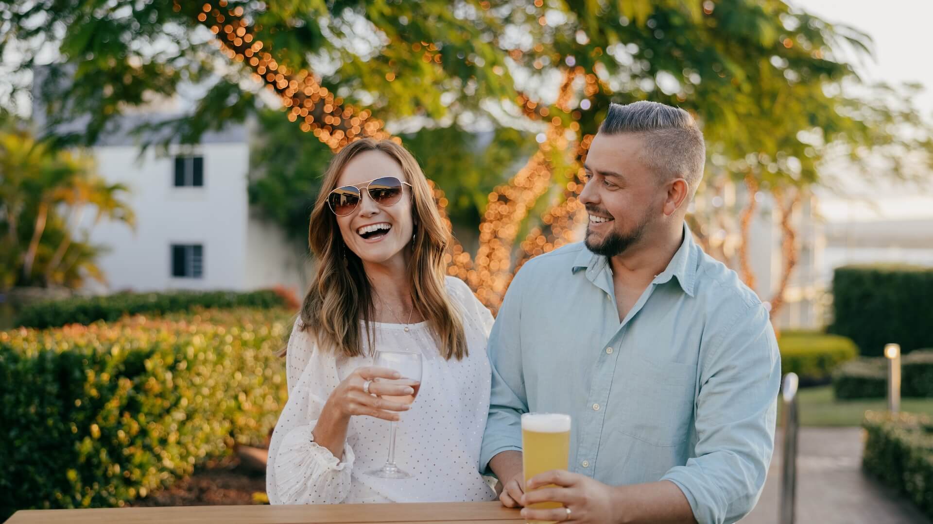 Two people smiling outdoors on the Lobby Deck, holding drinks. The woman wears sunglasses and a white dress, the man wears a light blue shirt. Trees with string lights in the background.