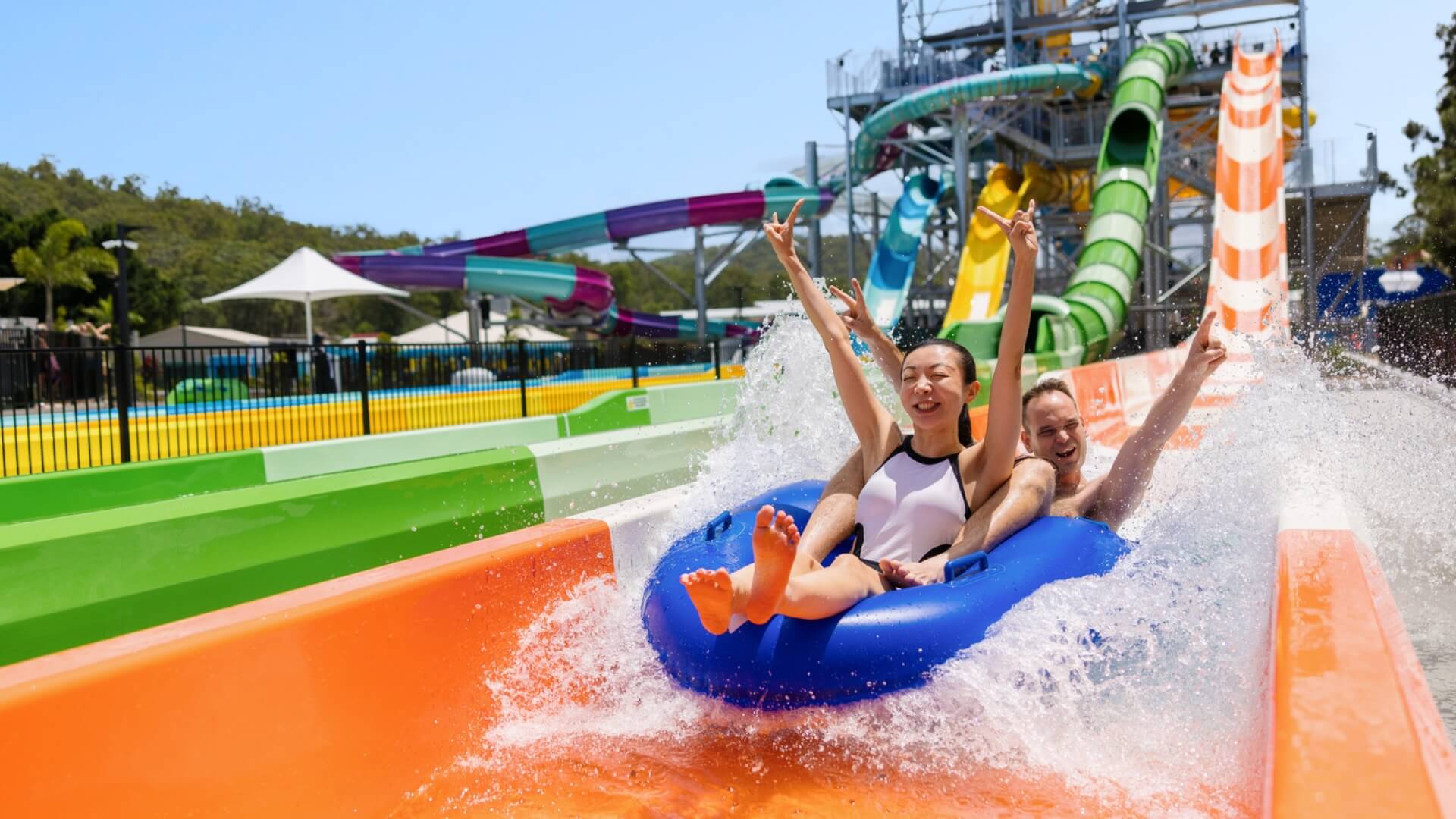 Two people ride a blue inflatable tube down Double Barrel water slide at Wet'n'Wild, surrounded by splashing water and vibrant slide structures in the background.