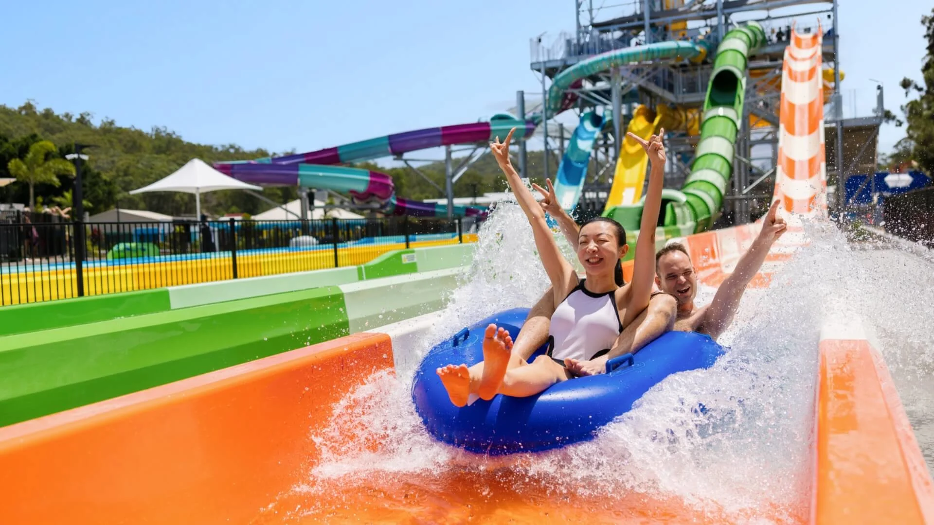 Two people ride a blue inflatable tube down Double Barrel water slide at Wet'n'Wild, surrounded by splashing water and vibrant slide structures in the background.