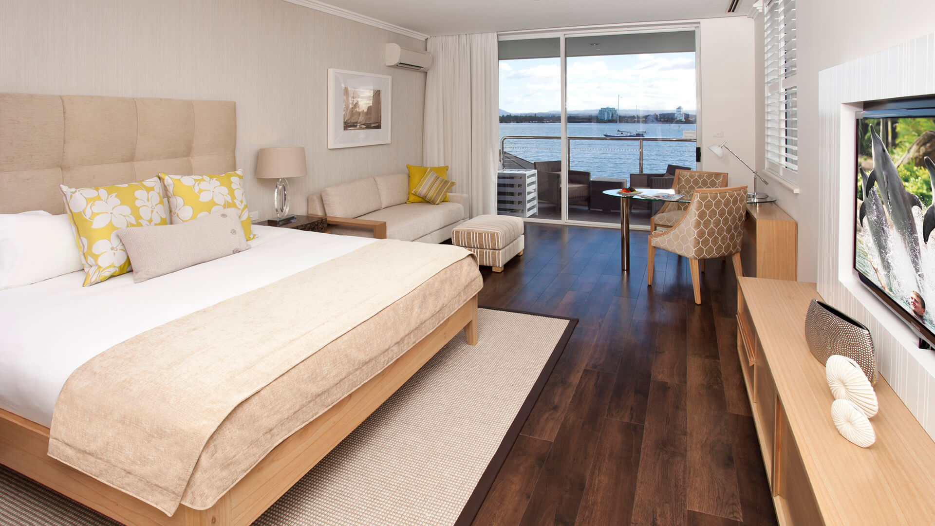 Hotel room with a large bed, a couch, a TV, and a small table. Sliding glass doors lead to a balcony with a view of water and boats. Room decor is in neutral tones with accent pillows.