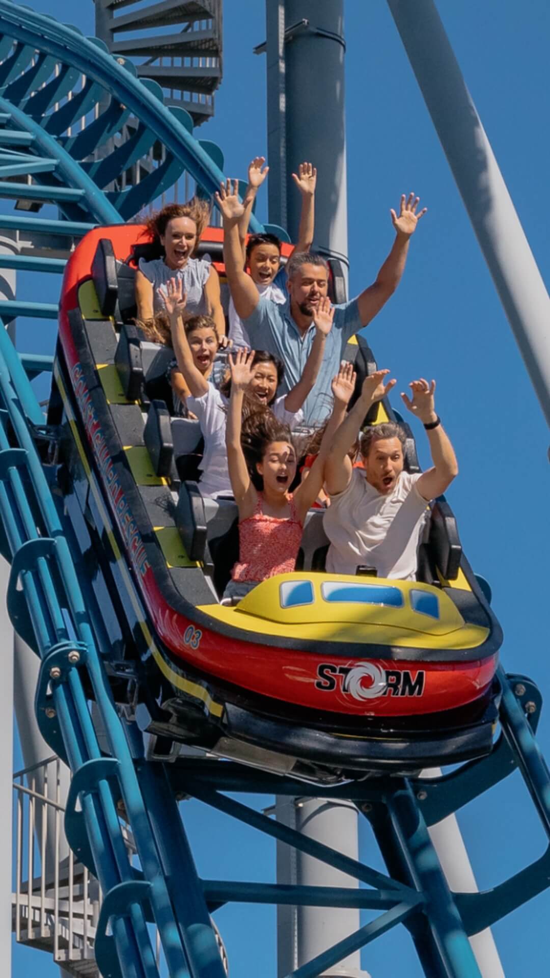 People are riding a colorful roller coaster with their hands up, enjoying the ride under a clear blue sky.