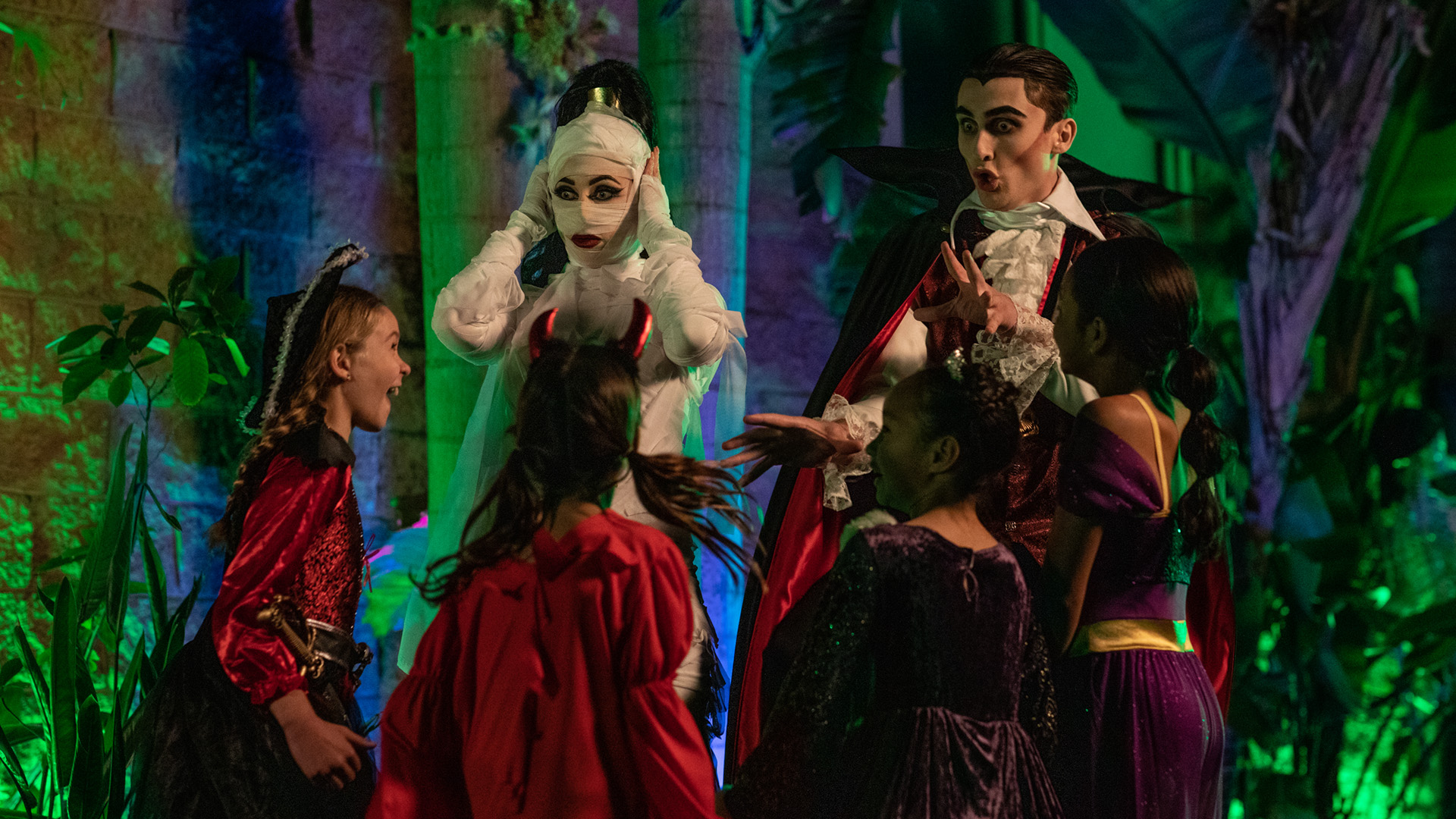 Five children in colorful Halloween costumes gather around two adults dressed as a ghost and a vampire, with lush green plants and dramatic lighting in the background. The ghost covers their ears while the vampire gestures animatedly.