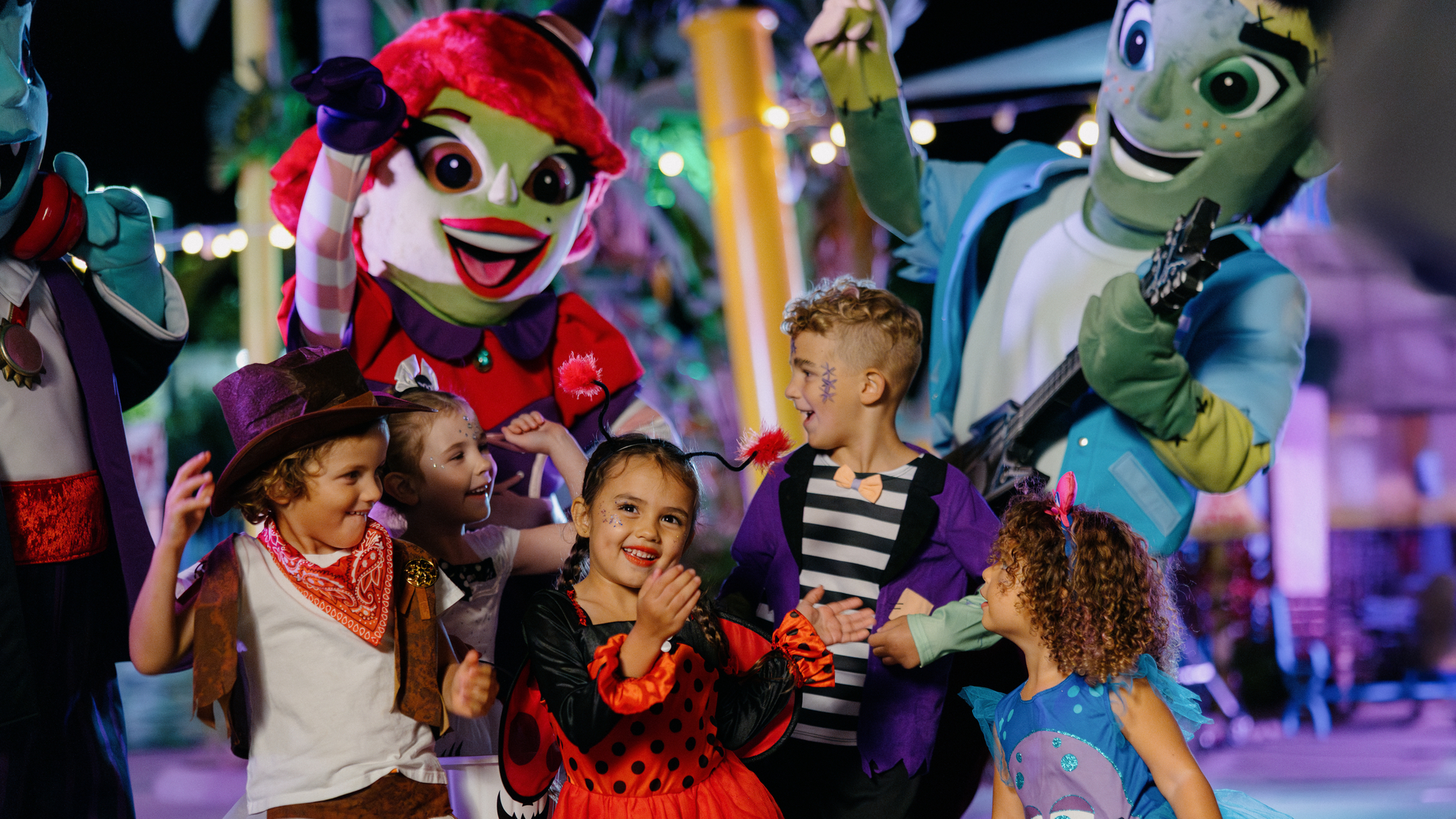 A group of children in Halloween costumes smile and pose with two colorful, friendly mascots at a lively outdoor event, with festive lights and decorations in the background.