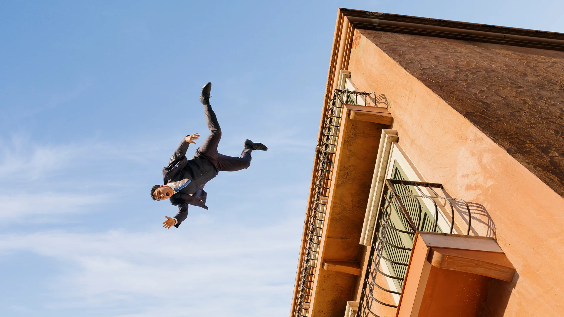 A man in a suit is mid-air, falling or jumping off the side of a peach-colored building against a blue sky. His arms and legs are spread out, and his expression looks surprised or alarmed.